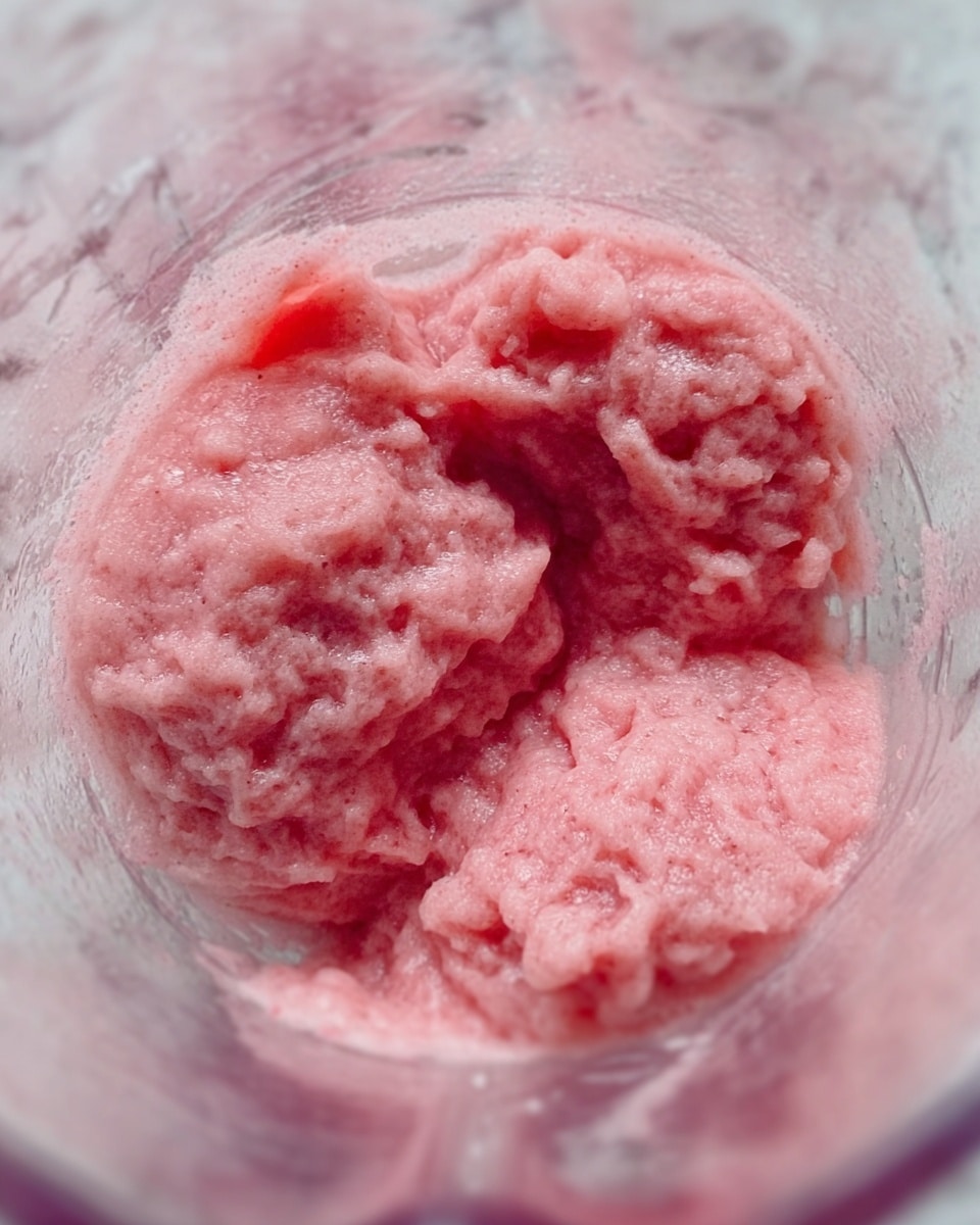 The image shows a close-up of three portions of raw pink ground meat inside a blender container. The texture looks soft and slightly chunky, with visible small meat pieces. The blender has a transparent body, and the meat’s pink color contrasts with the clear container. The background is a white marbled texture. photo taken with an iphone --ar 4:5 --v 7