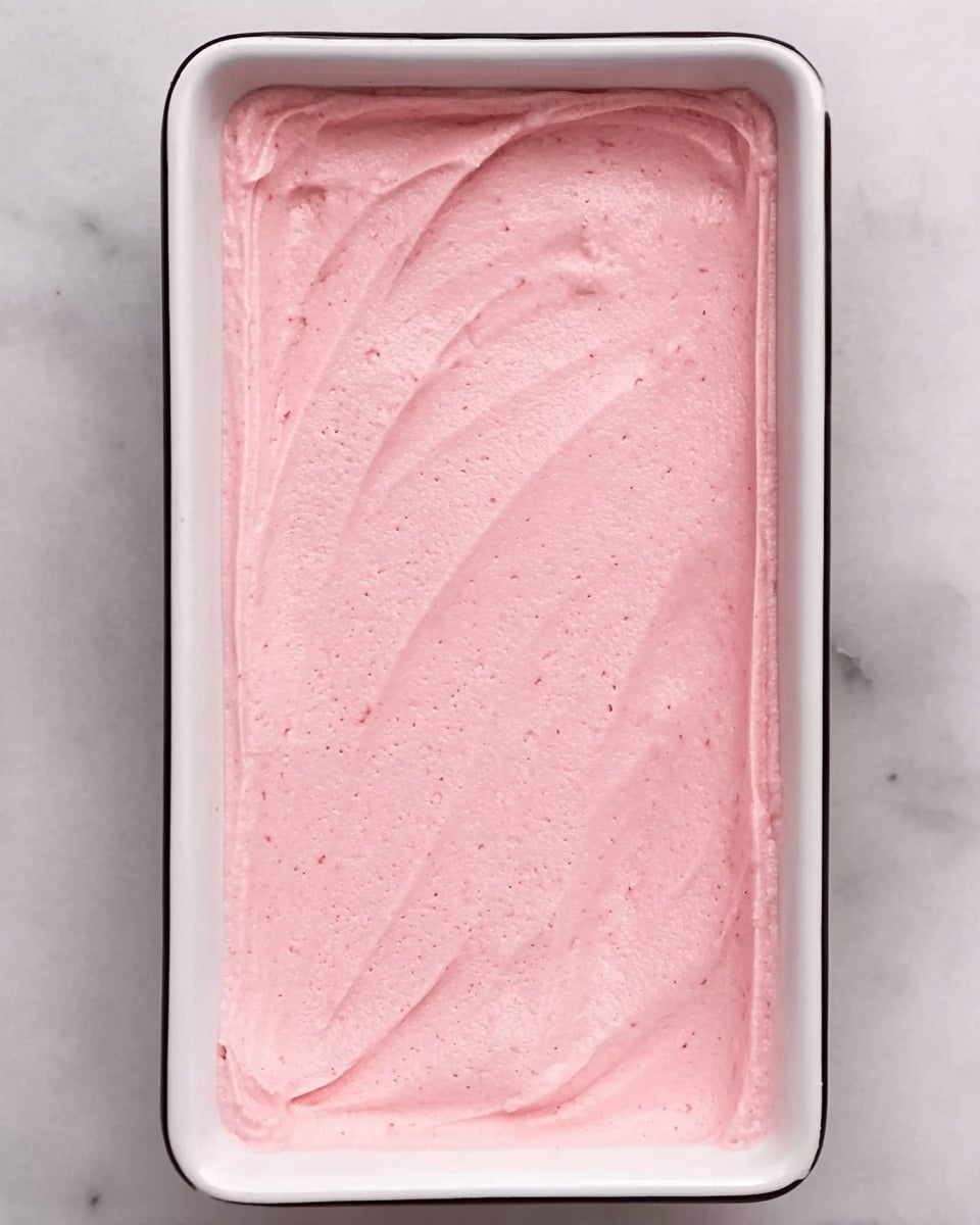The image shows a shallow rectangular white dish filled with a smooth, pink creamy mixture spread evenly across the surface. The texture looks soft and slightly fluffy with gentle, uneven lines created by a spreading tool on the top. The background is a white marbled surface. photo taken with an iphone --ar 4:5 --v 7