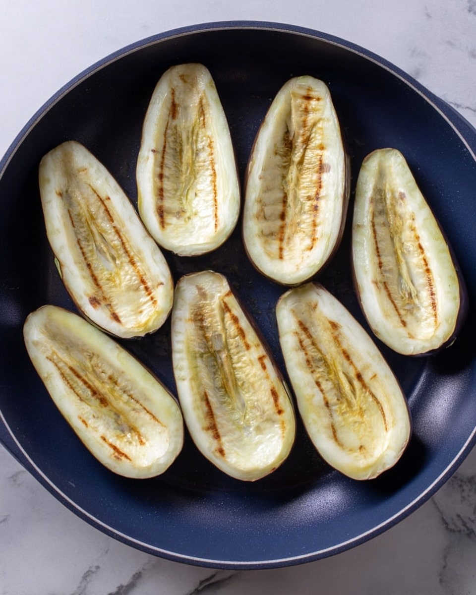 The image shows eight halves of peeled eggplants arranged in a single layer inside a round dark blue pan. Each eggplant half has a light cream color with patches of golden brown grill marks and a soft texture on the surface. The pan is placed on a white marbled surface that contrasts with the darker pan and pale eggplants. The eggplants are evenly spaced, filling the pan without overlapping. photo taken with an iphone --ar 4:5 --v 7
