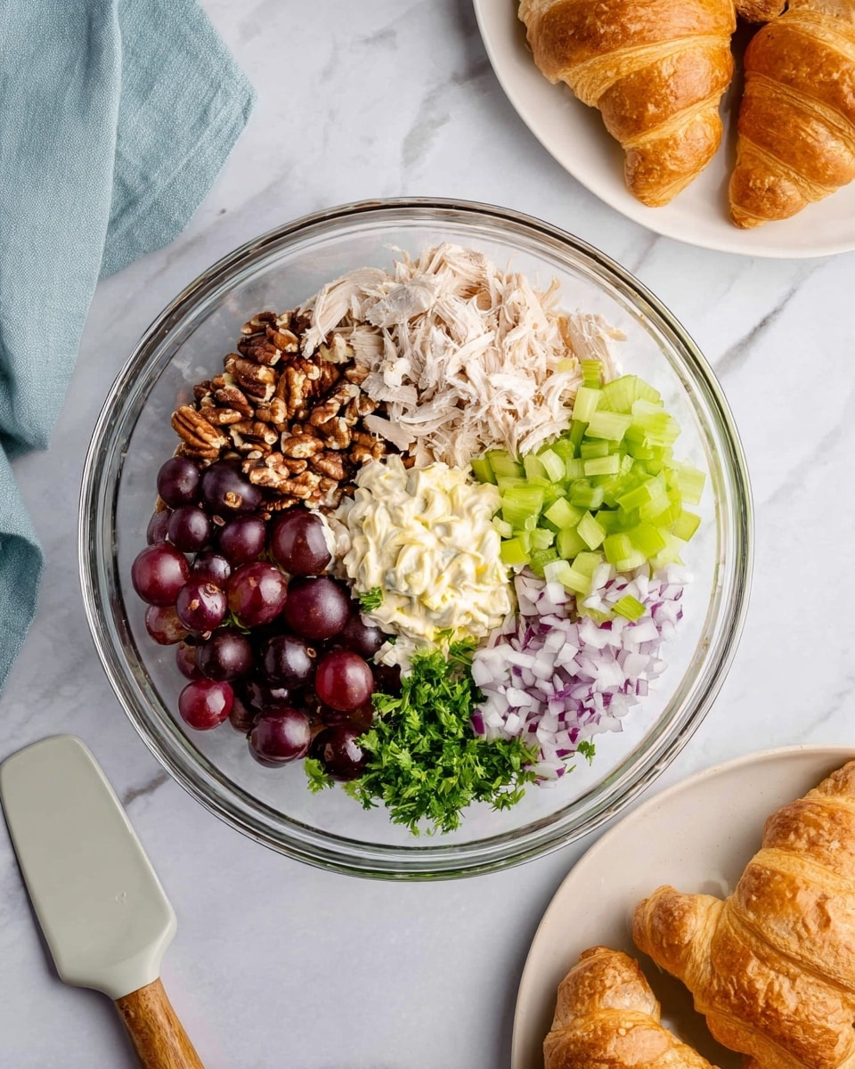 A clear glass bowl on a white marbled surface holds six separate piles of ingredients arranged in sections: dark red grapes cut in halves at the bottom left, chopped brown pecans at the top left, shredded light beige chicken in the center, chopped purple onions at the top right, bright green chopped celery at the middle right, a dollop of pale yellow mayonnaise near the bottom center, and finely chopped dark green parsley at the bottom edge. Nearby, a white plate holds four golden brown croissants with flaky texture. A light gray spatula and a light blue cloth are also on the surface. Photo taken with an iphone --ar 4:5 --v 7