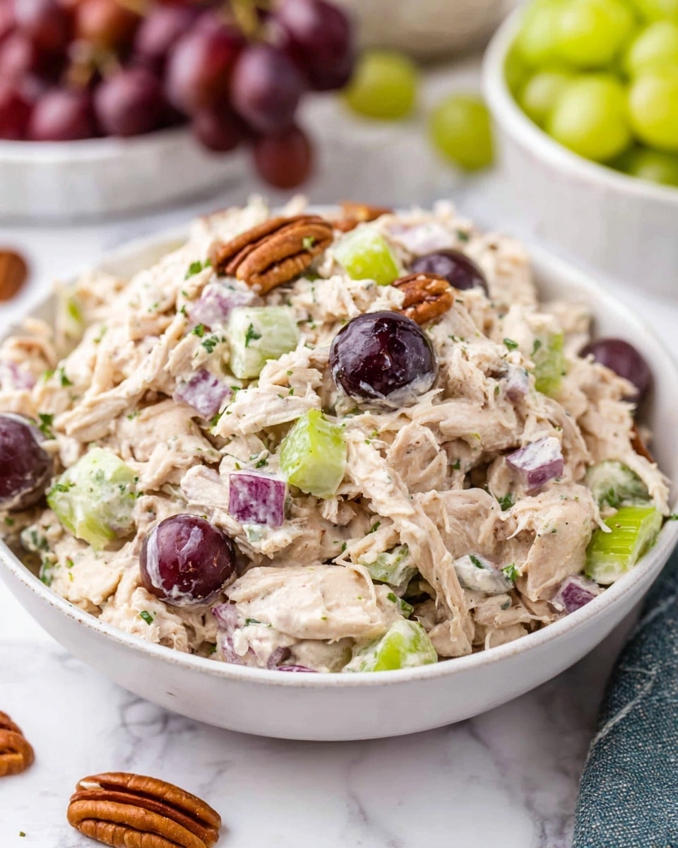A close-up view of a creamy chicken salad served in a white bowl placed on a white marbled surface, showing a mix of shredded chicken mixed with a light beige creamy dressing, chopped light green celery pieces, small purple grape halves, and bits of red onion with tiny green herb sprinkles spread throughout. The salad also contains halved dark purple grapes and small crunchy pecan pieces scattered inside and outside the bowl, with whole pecans placed nearby on the surface. In the background, other white bowls with green grapes and red grapes are blurred, adding soft color contrast to the scene. Photo taken with an iphone --ar 4:5 --v 7