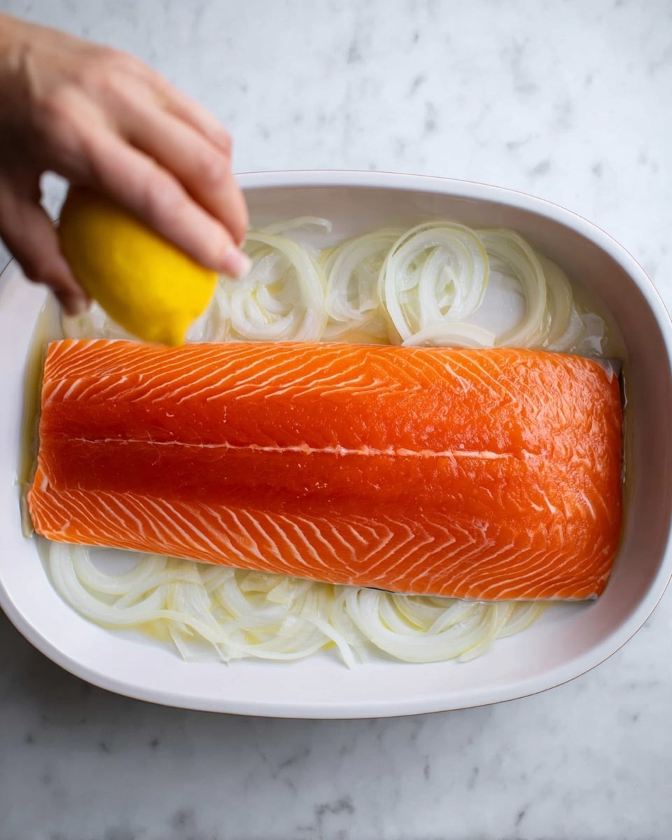 A large, bright orange raw salmon fillet lies flat on a bed of thin, white onion slices in a white oval baking dish. The salmon shows detailed, natural lines across its smooth surface, while the onion slices beneath create a soft, layered background. A woman's hand is squeezing a fresh, yellow lemon half over the salmon from the lower right corner, adding a fresh and bright element to the scene. The dish is placed on a white marbled surface that provides a clean and simple backdrop, highlighting the vivid colors of the ingredients photo taken with an iphone --ar 4:5 --v 7