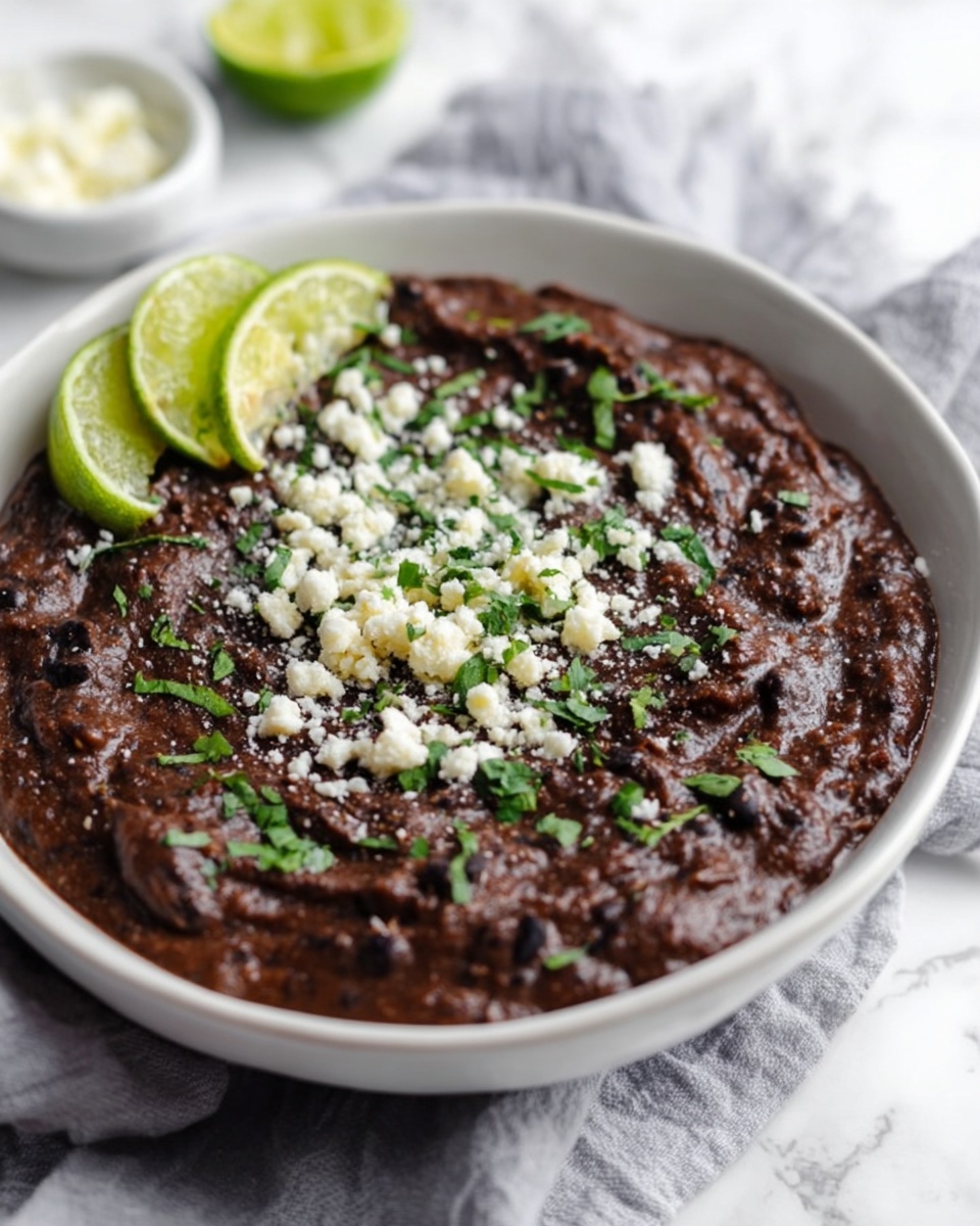 A white bowl filled with a thick, dark brown layer of refried black beans, topped with small white crumbles of cheese and chopped green herbs scattered mainly in the center and edges, with two wedges of bright green lime placed on the rim of the bowl at the top; the bowl sits on a light gray cloth over a white marbled surface, with a blurred white bowl and lime wedge in the background, photo taken with an iphone --ar 4:5 --v 7