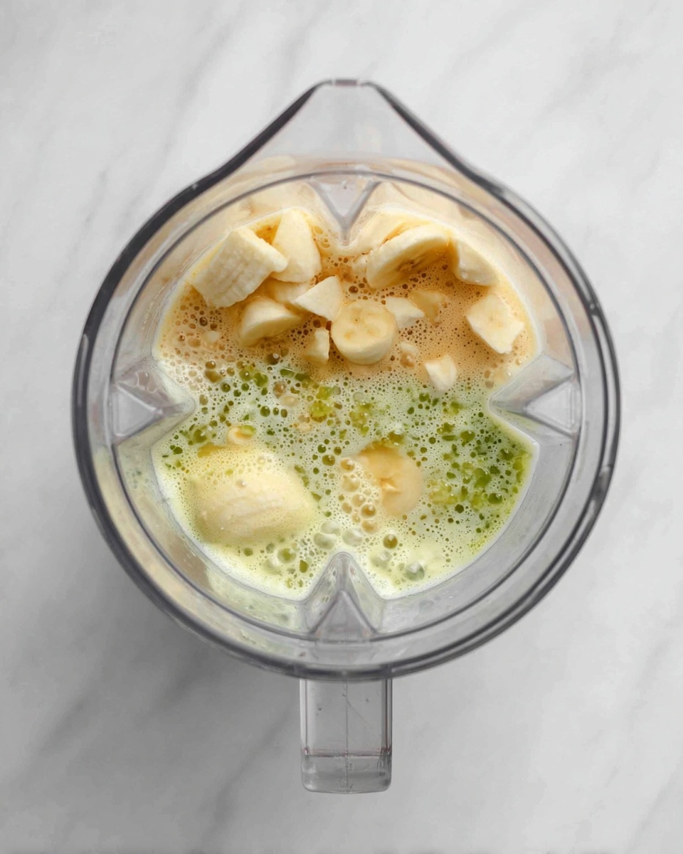 A top view of a clear blender with three layers inside: at the bottom, a creamy light beige layer; in the middle, a bubbly, bright green layer with small bits giving a textured look; on top, uneven chunks of pale yellow banana pieces scattered around. The blender is placed on a white marbled surface. photo taken with an iphone --ar 4:5 --v 7