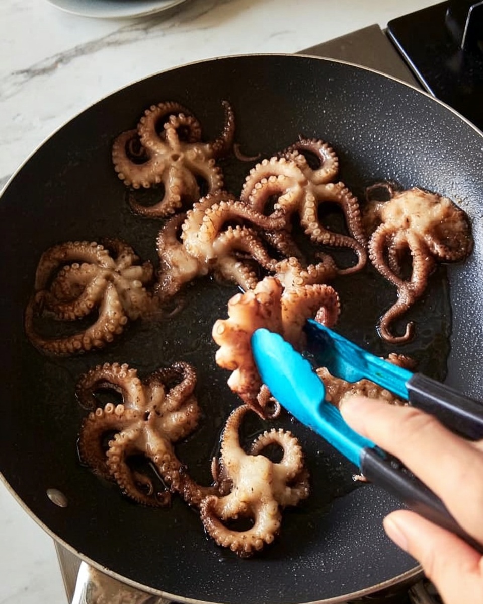A black pan holds several small octopuses being cooked. The octopuses have light beige and brown colors, showing a cooked, slightly crispy texture on their tentacles and heads. A woman's hand is using blue tongs to lift one of the octopuses in the pan. The background and surface nearby have a white marbled texture. Photo taken with an iphone --ar 4:5 --v 7