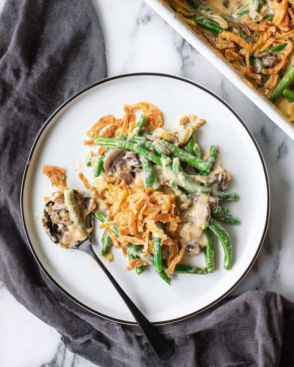 A white plate with a thin black rim holds one serving of green bean casserole. The dish has three visible layers: fresh green beans that are bright green and firm, a creamy light beige sauce with small mushroom pieces mixed inside, and a top layer of crispy golden fried onions scattered unevenly over the beans and sauce. Next to the plate is a silver spoon with some of the casserole on it, showing the same layers, and a black fork rests on the edge of the plate. The plate sits on a white marbled surface with a dark gray cloth nearby. In the upper right corner, there is a white rectangular baking dish filled with more of the same casserole. Photo taken with an iphone --ar 4:5 --v 7