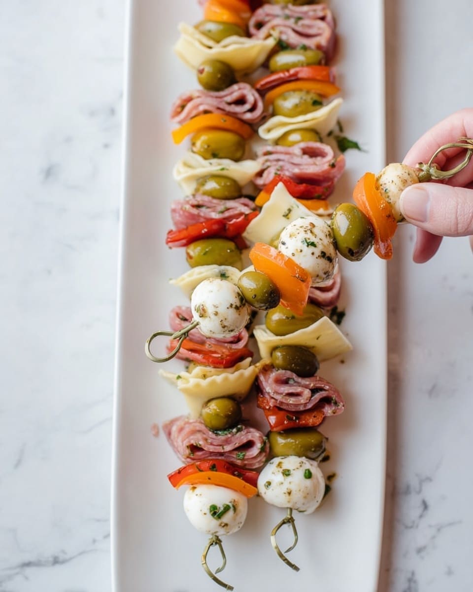 Several skewers are arranged on a long white plate placed on a white marbled surface. Each skewer has five layers from right to left: a green olive with seasoning, a folded slice of pink salami, a piece of pale yellow cheese folded like a small ravioli, a bright orange bell pepper slice, and a small white round mozzarella ball with herbs. A woman's hand is holding one skewer by the looped end. Photo taken with an iphone --ar 4:5 --v 7