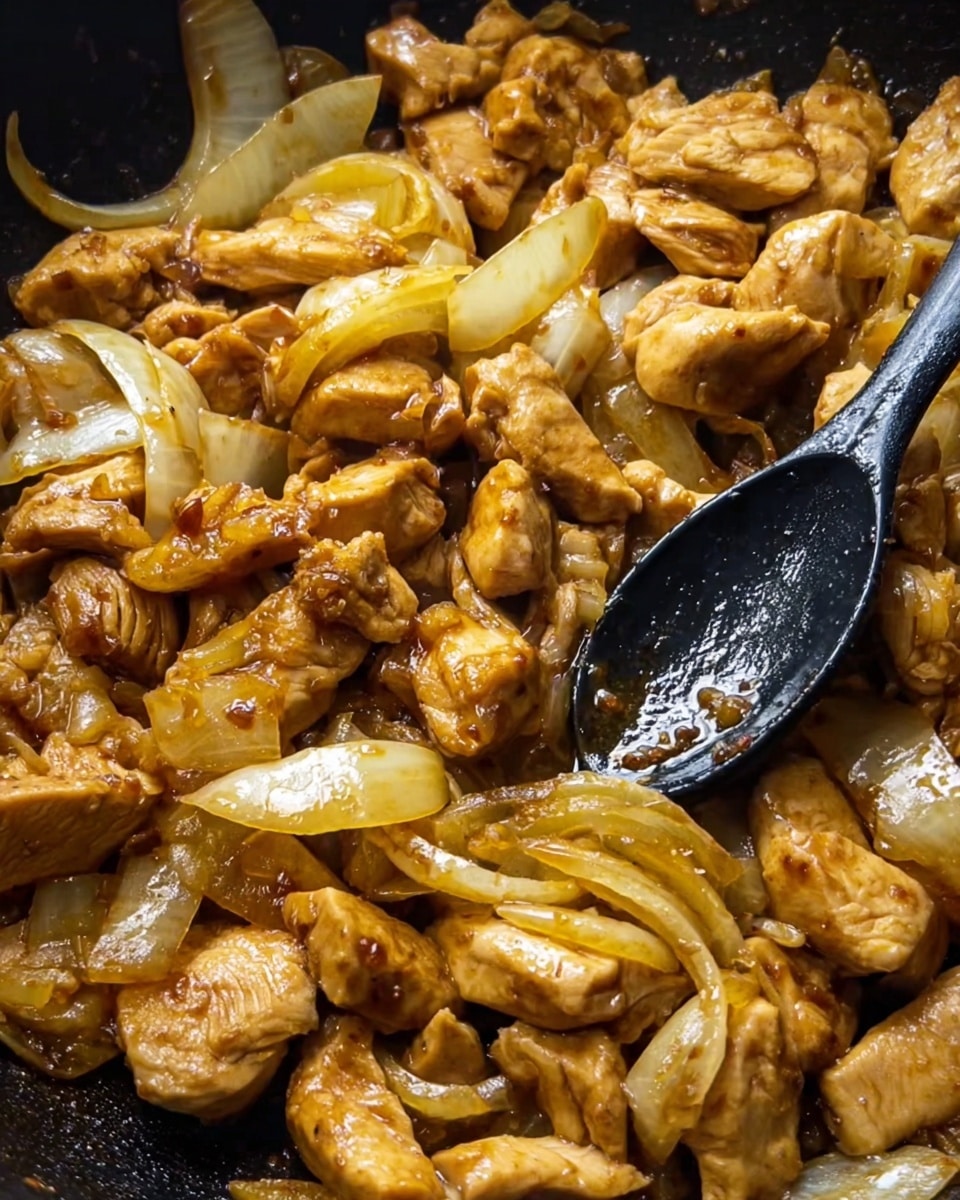 The image shows a close-up of many pieces of cooked chicken mixed with sliced onions in a pan. The chicken pieces are golden brown with a shiny, slightly saucy texture. The onions, cut into thick pieces, are translucent and yellowish, spread evenly among the chicken. A black cooking spoon lies on top of the mixture with a little sauce on it. The background is dark inside the pan, focusing on the cooked food, with no other items visible. Photo taken with an iphone --ar 4:5 --v 7