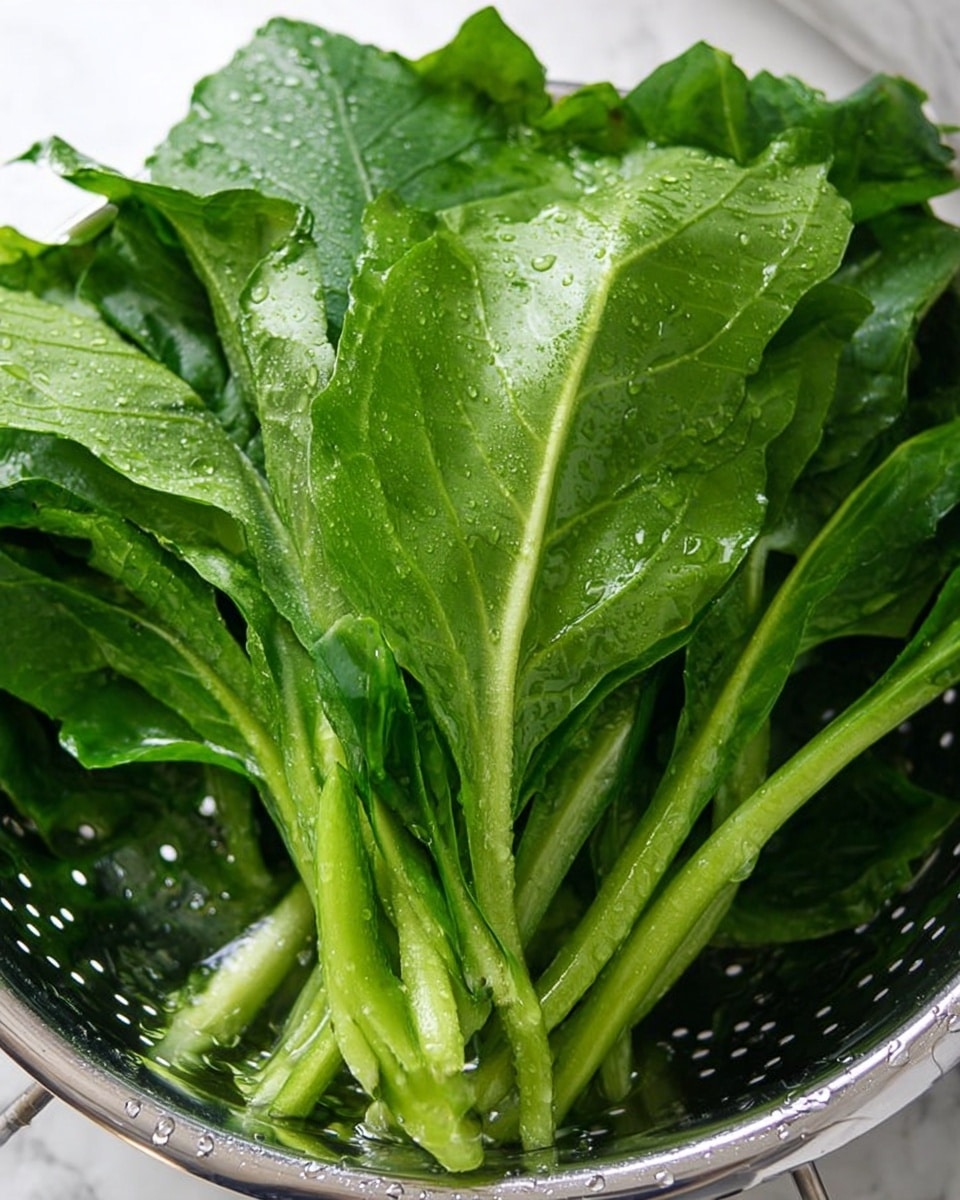 The image shows a close-up of fresh green leaves in a silver colander. The leaves are large with smooth and slightly shiny surfaces, showing some water droplets on them. Their stems are long and light green, lying across the pile of leaves. The colander's edge is visible, shining with water drops, and the whole setup sits on a white marbled texture. The leaves look fresh and crisp, filling the colander fully. photo taken with an iphone --ar 4:5 --v 7