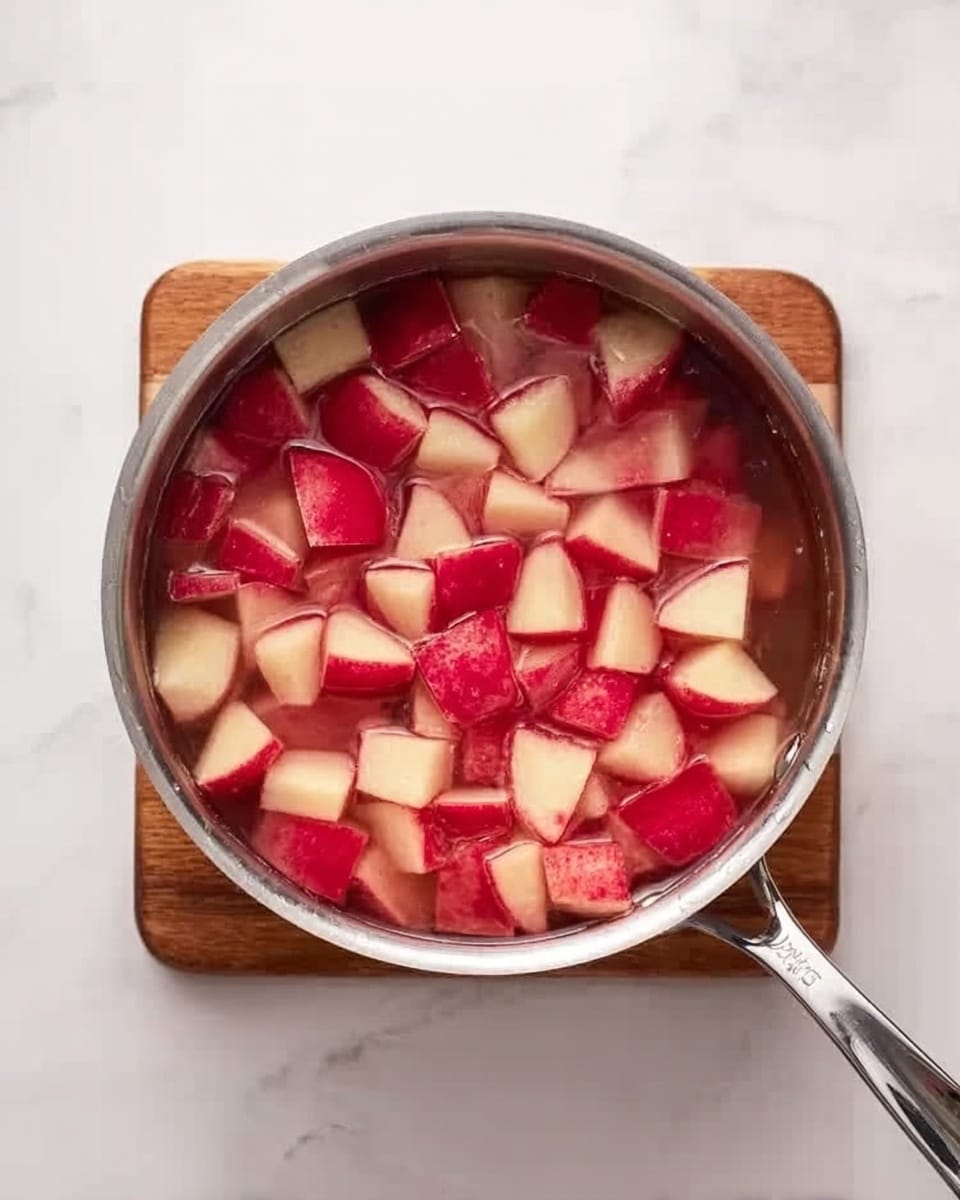 The image shows a silver pot filled with cubed red-skinned potatoes submerged in water. The potatoes are evenly cut into medium-sized pieces and float within the clear liquid, with some pieces touching the sides of the pot. The pot sits on a small wooden board placed on a white marbled surface. The pot handle extends to the right side of the frame. Photo taken with an iphone --ar 4:5 --v 7