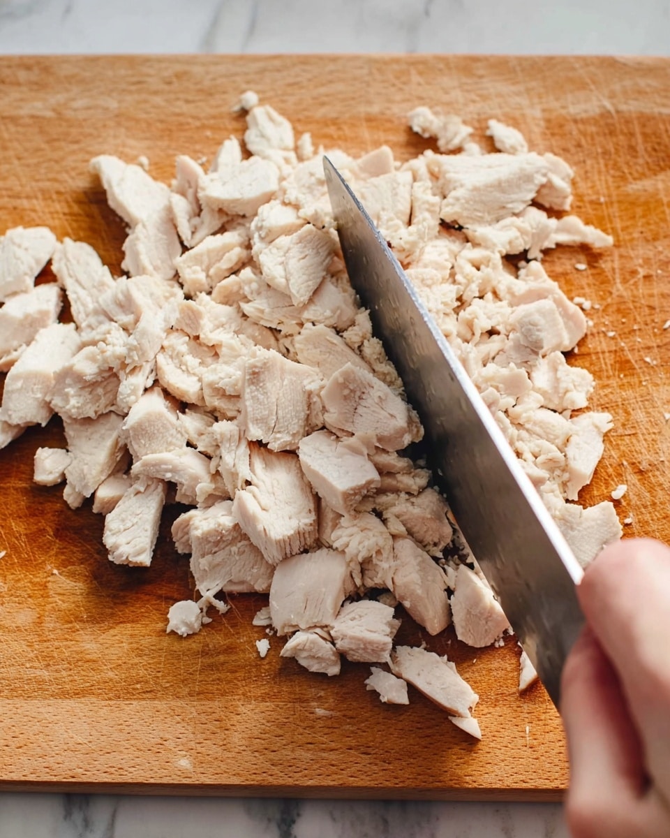 A close-up view of a wooden cutting board with many small white pieces of cooked chicken scattered across its surface. A metal knife is positioned in the middle, slicing through the chicken pieces, with a woman's hand holding the knife on the right side of the frame. The wooden cutting board has a warm light brown color with visible grain texture. The background shows a white marbled texture. photo taken with an iphone --ar 4:5 --v 7