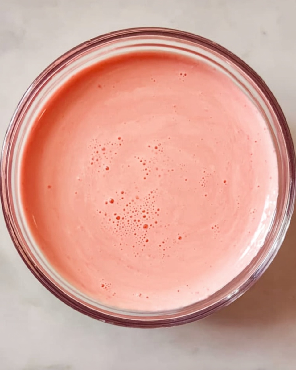 A clear glass bowl filled with smooth, creamy light pink sauce with a few small bubbles on the surface, set on a white marbled surface. The sauce has a consistent texture and a gentle swirl pattern inside the bowl. Photo taken with an iphone --ar 4:5 --v 7