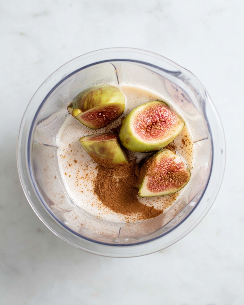 A clear blender container sits on a white marbled surface, inside there are five fig pieces with green outer skin and pinkish-red inner flesh. The figs rest in a small pool of creamy liquid with some foam on top. A line of light brown cinnamon powder curves along one inner side of the container, contrasting with the creamy white liquid and the figs. The textures are smooth and fresh, with the figs showing a soft and juicy look. photo taken with an iphone --ar 4:5 --v 7