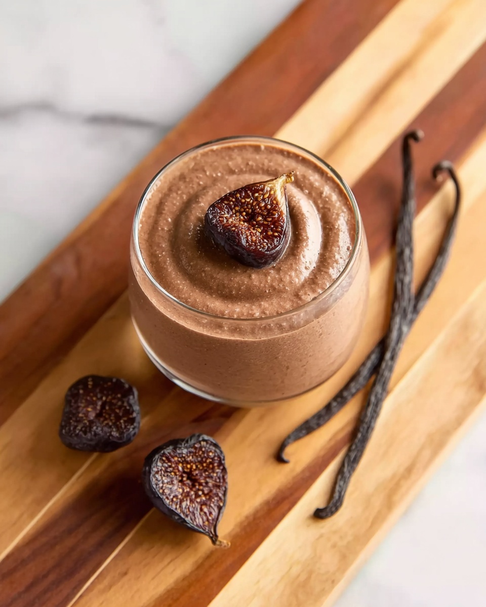 A glass cup filled with a thick, smooth brown mousse topped with a small dried fig piece in the center. The cup sits on a wooden board with visible light and dark stripes. There are dried figs and two long vanilla pods placed around the glass on the wooden board. The background is a white marbled surface. photo taken with an iphone --ar 4:5 --v 7
