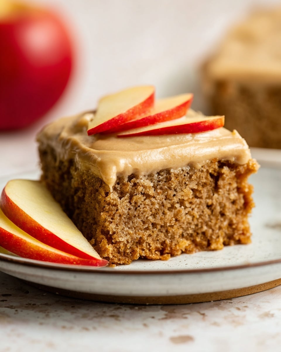 The image shows a square piece of moist brown cake with a crumbly texture on a white plate with a thin rim. The cake has one thick layer topped by a smooth, light brown frosting layer. Two thin slices of shiny red and yellow apple rest on top of the frosting, and a few more apple slices lean against the cake on the plate. The background is a soft white marbled texture, with a blurred red apple visible in the back. Photo taken with an iphone --ar 4:5 --v 7