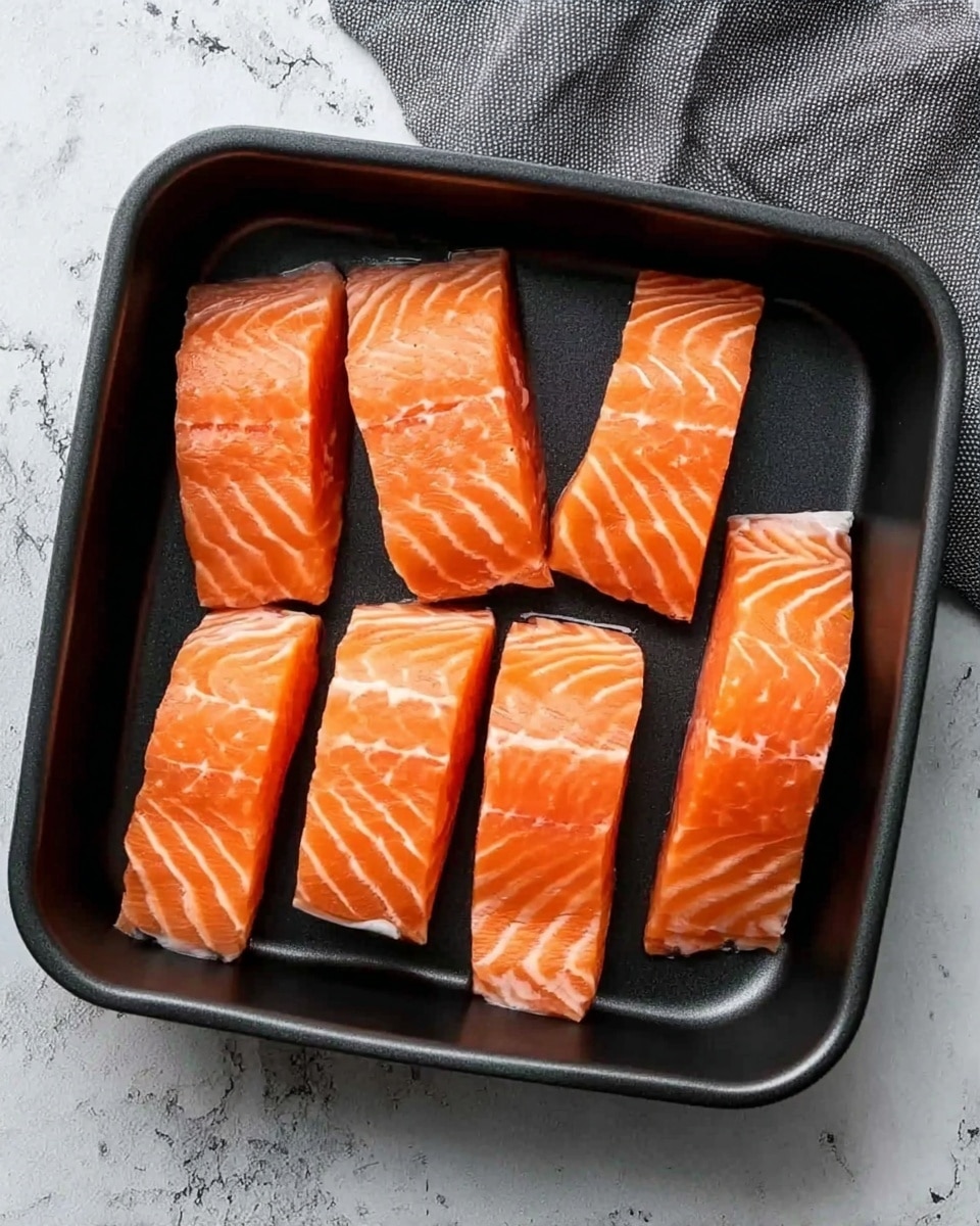 The image shows seven pieces of raw salmon arranged neatly in a black square pan. The salmon pieces are bright orange with white lines running through them, showing fresh texture. There are two shorter pieces in the middle column and five longer pieces around them. The pan is placed on a surface with a white marbled texture, next to a grey cloth. The overall look is clean and fresh, with the salmon pieces clearly standing out. photo taken with an iphone --ar 4:5 --v 7