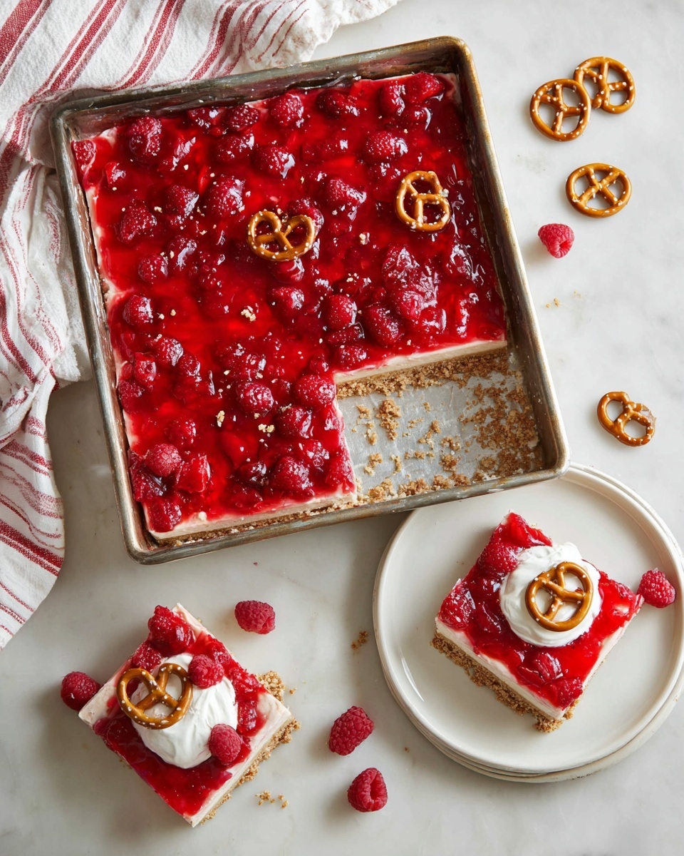 The image shows a square dessert bars with three visible layers placed in a rectangular metal baking pan on a white marbled surface. The bottom layer is a crumbly tan crust, the middle layer is a light cream cheese or similar spread, and the top layer is a thick glossy bright red fruit topping with whole and sliced raspberries. One bar has been removed from the pan and is placed on a white plate to the right, showing all three layers. Another bar is on a round white plate at the bottom, topped with a dollop of white cream and a small pretzel. Several small pretzels are scattered around the pan and plates. A white cloth with red stripes is under the baking pan and near the plate on the right. Photo taken with an iphone --ar 4:5 --v 7