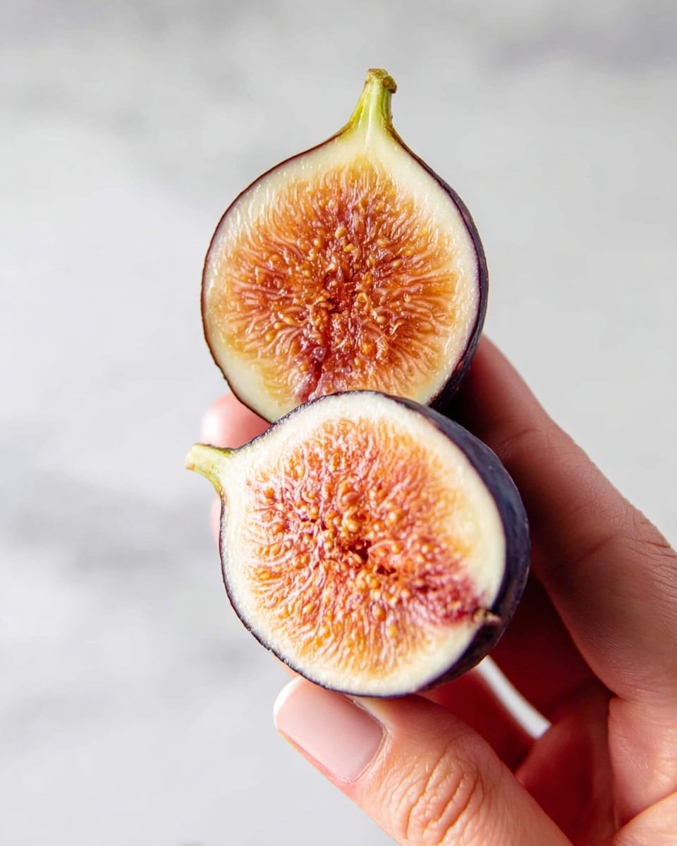 A close-up image shows a woman's hand holding two halves of a ripe fig, split vertically. The fig's thin outer skin is dark purple, with a green stem at the top. Inside, the fruit reveals a soft, juicy texture in shades of orange and pink, with small seeds visible throughout the flesh. The background is a white marbled texture. photo taken with an iphone --ar 4:5 --v 7