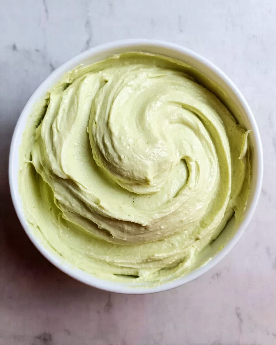 A white bowl filled with a single creamy pale green layer that has a smooth and whipped texture, with soft swirls on the surface showing gentle peaks and valleys. The bowl sits on a white marbled surface. Photo taken with an iphone --ar 4:5 --v 7