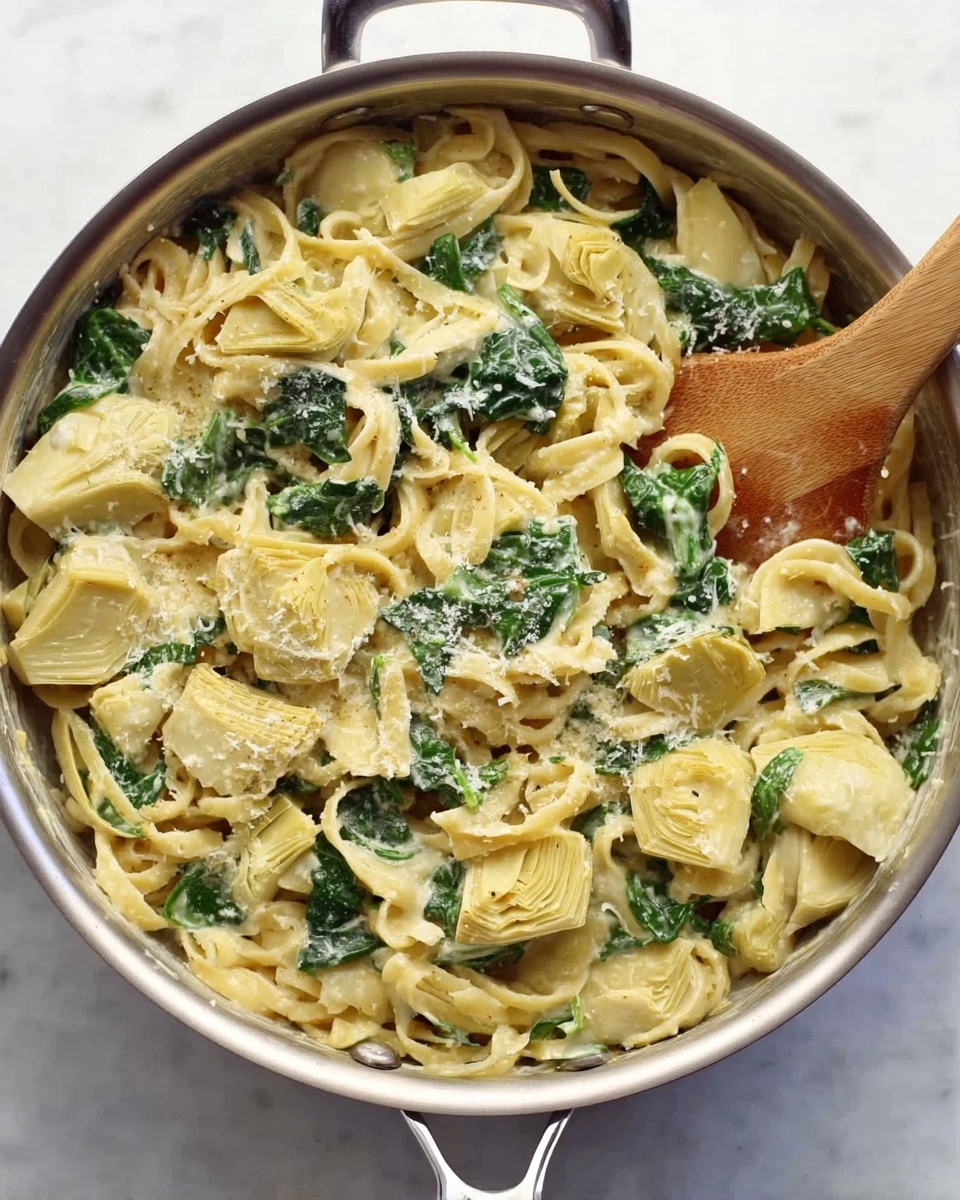 A silver pan filled with creamy pasta mixed with light yellow artichoke hearts and dark green spinach leaves. The pasta is coated in a pale yellow sauce with a slightly grainy texture, sprinkled with grated cheese. A wooden spoon rests on the side inside the pan. The pan sits on a white marbled surface. photo taken with an iphone --ar 4:5 --v 7