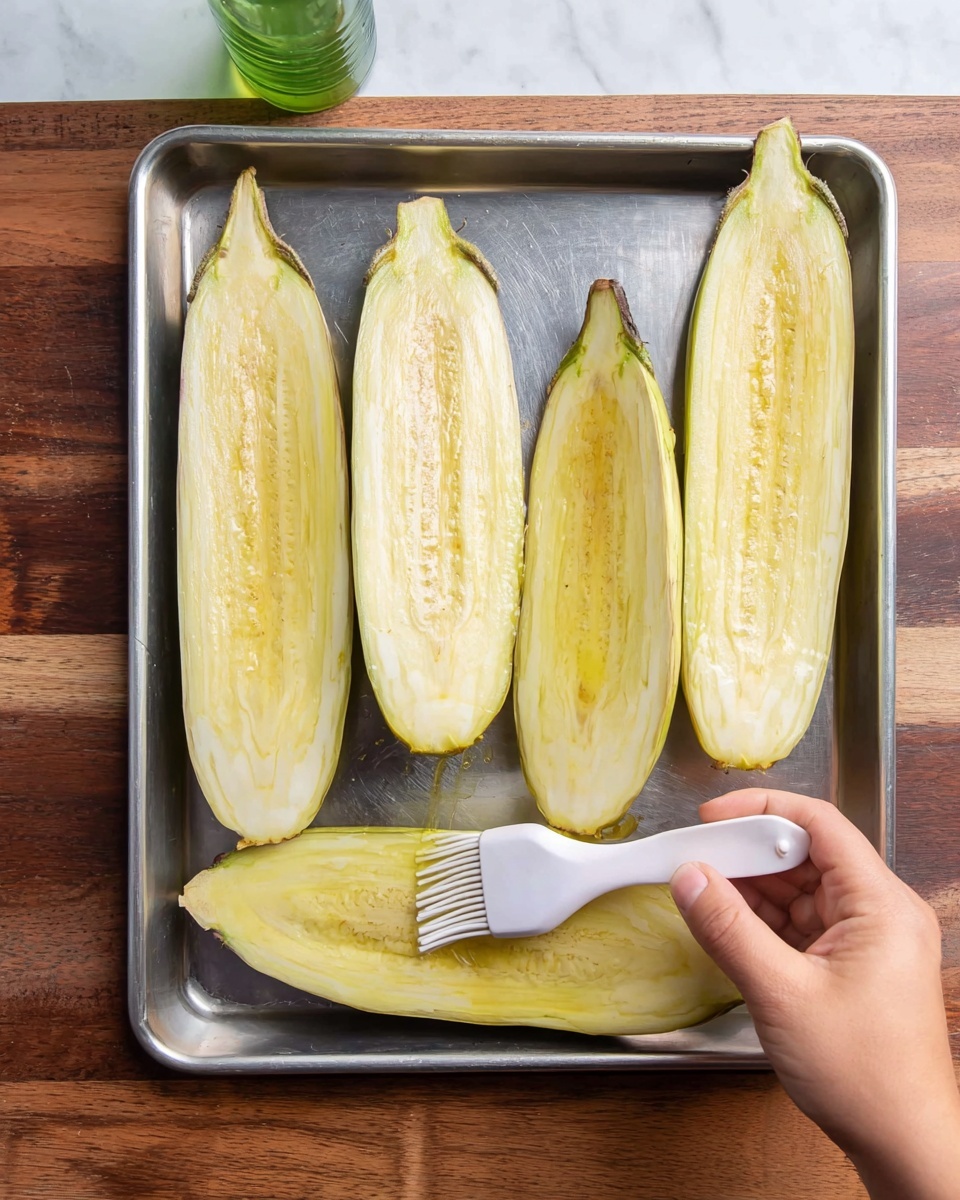Four eggplants cut in half lengthwise lie on a metal baking tray, placed on a wooden surface now described as a white marbled texture. The eggplant halves show a pale yellow inside with light brown seed spots and a smooth wet texture. A woman's hand holds one eggplant half while brushing oil onto its surface with a white silicone brush. A small green bottle is seen in the upper left corner. photo taken with an iphone --ar 4:5 --v 7