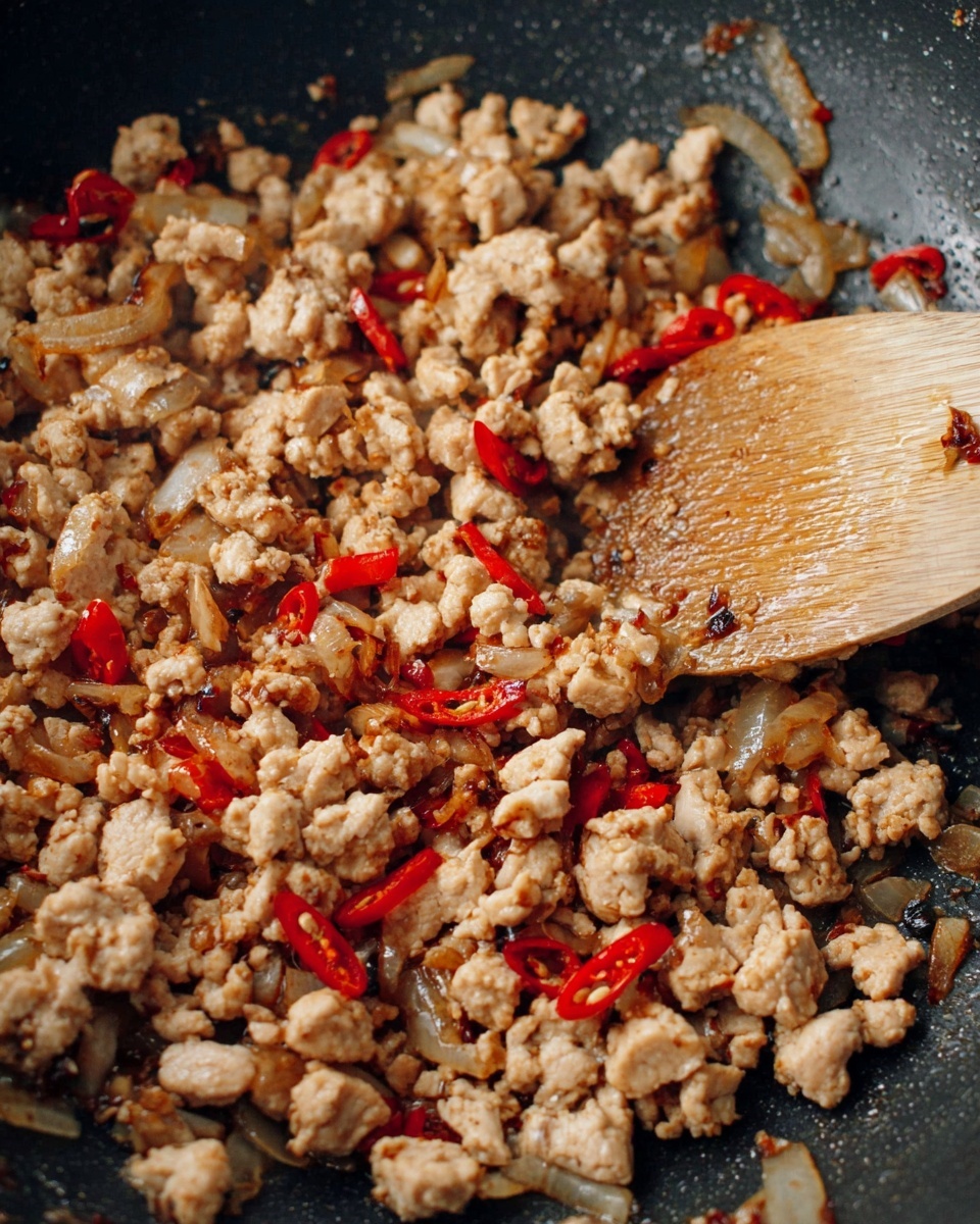 The image shows a skillet filled with small pieces of cooked minced meat mixed with thin slices of red chili and translucent onion. The meat has a light brown color with slight browning and some darker, caramelized bits scattered around. The red chili slices add bright red spots throughout the dish, and the onions are slightly caramelized with a soft brown color. A wooden spoon is resting on top of the mixture, partly covered in bits of the cooked food. The skillet’s dark surface contrasts with the light colors of the meat and vegetables. The photo taken with an iphone --ar 4:5 --v 7