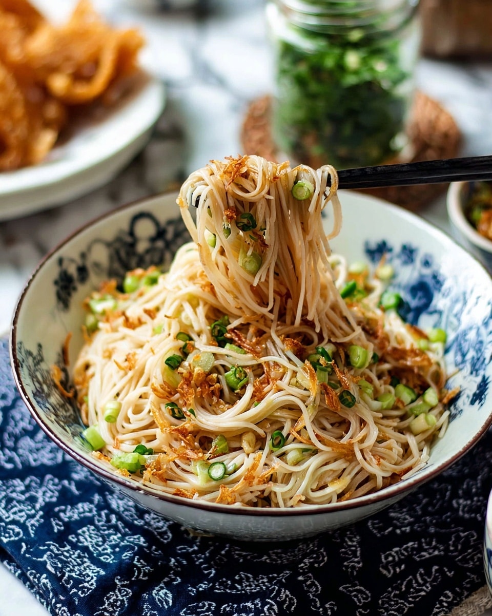 A bowl filled with a nest of thin, light beige noodles, layered with small pieces of green onion and some golden brown fried shallots scattered on top and mixed throughout. A pair of black chopsticks holds a bunch of noodles lifted gently from the bowl. The bowl is white with a blue floral pattern inside, and it sits on a dark blue patterned cloth over a white marbled surface. In the background, there is a blurred white plate with crispy golden brown pieces and a glass jar with green herbs. Photo taken with an iphone --ar 4:5 --v 7
