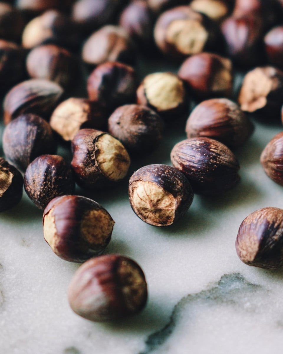 The image shows many hazelnuts with dark brown shells and some light tan areas where the shells have cracked open, spread out on a flat surface with a white marbled texture in the background. The nuts vary in size and have a rough texture on their shells. The focus is on the middle area with hazelnuts close together, while the nuts farther from the center appear softly blurred. photo taken with an iphone --ar 4:5 --v 7