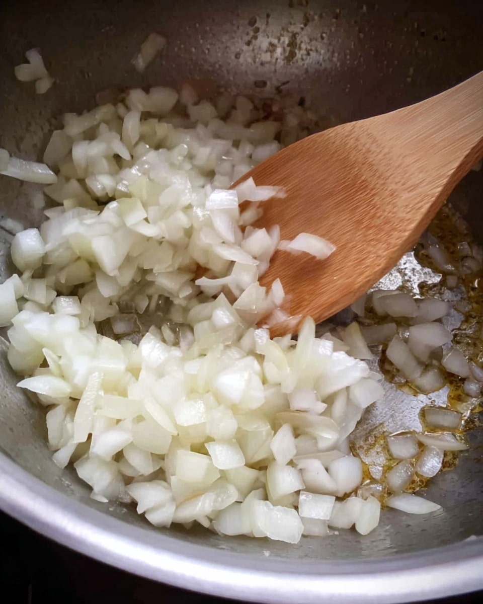 Inside a metal pot, small chopped onion pieces are being stirred with a wooden spoon. The onions are pale white with some parts turning translucent as they cook. The pot surface shows some light browning from cooking, giving it a slightly shiny texture. The wooden spoon is light brown and positioned at the top part of the pot, moving the onions gently. photo taken with an iphone --ar 4:5 --v 7