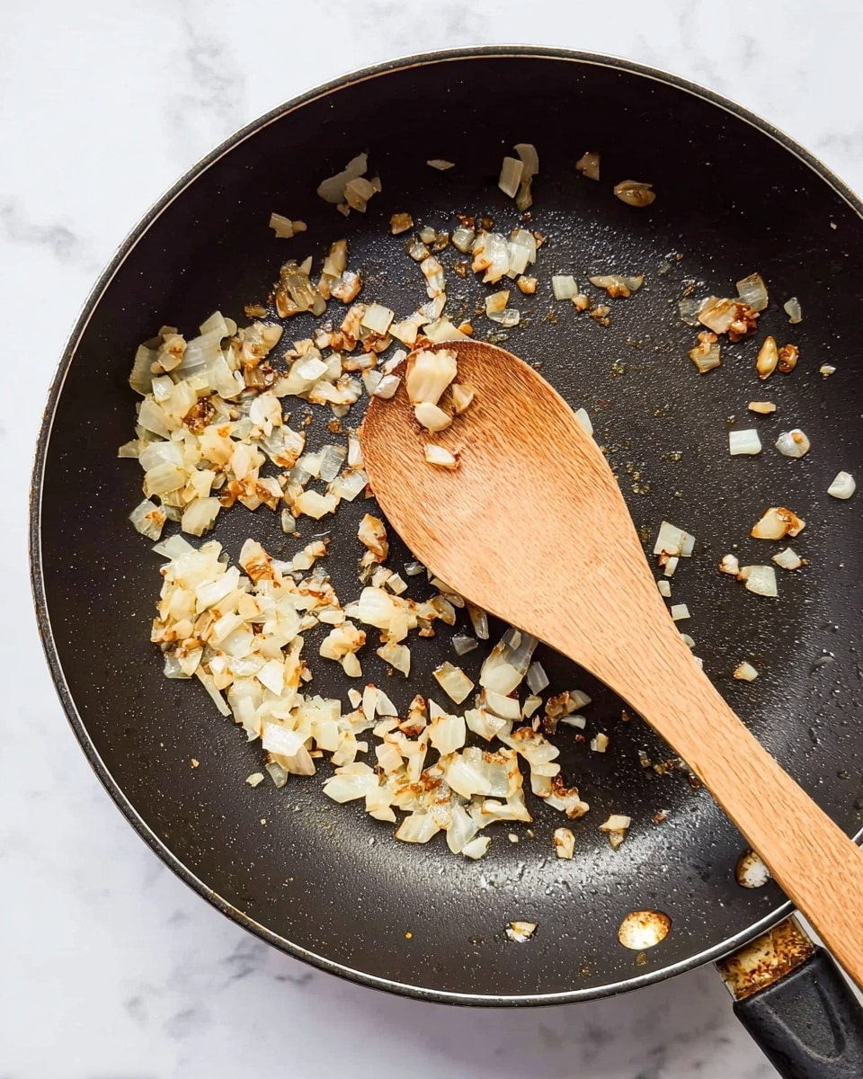 A black frying pan with some cooked, small pieces of onion and garlic that are light brown and translucent, scattered unevenly across the pan's bottom. A wooden spoon with a smooth texture lies inside the pan, resting diagonally from the top right to the lower left, with some bits of cooked onion sticking to its surface. The pan itself shows signs of use with some slight rust around the edges. The background is a white marbled surface. photo taken with an iphone --ar 4:5 --v 7