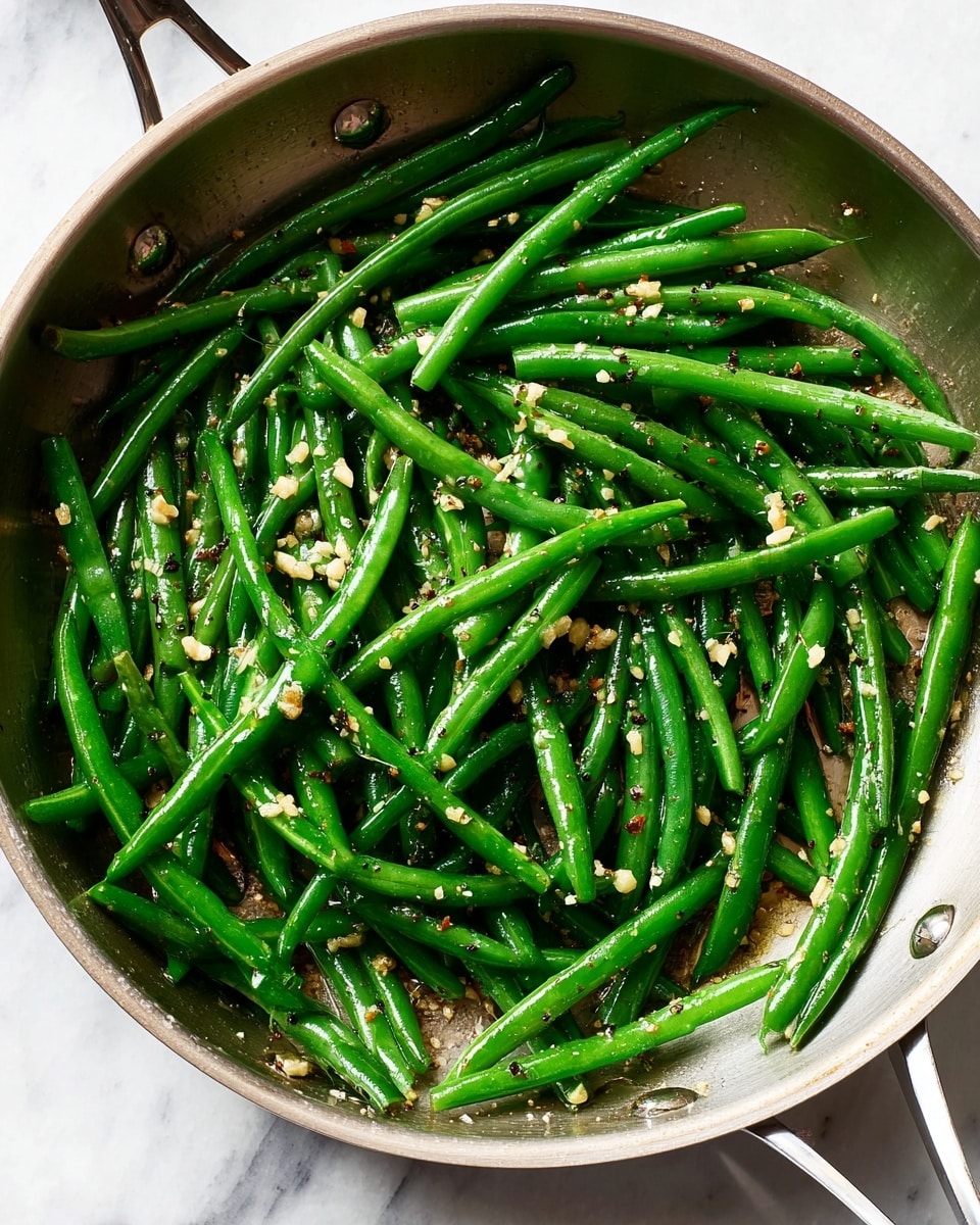 A stainless steel pan filled with bright green cooked green beans spread evenly, topped with small bits of light beige garlic and coarse black pepper flakes scattered throughout. The pan handle is visible at the top, and the surface underneath the pan is a white marbled texture. The green beans have a shiny texture from being lightly cooked with oil and seasonings, showing a fresh and simple look photo taken with an iphone --ar 4:5 --v 7