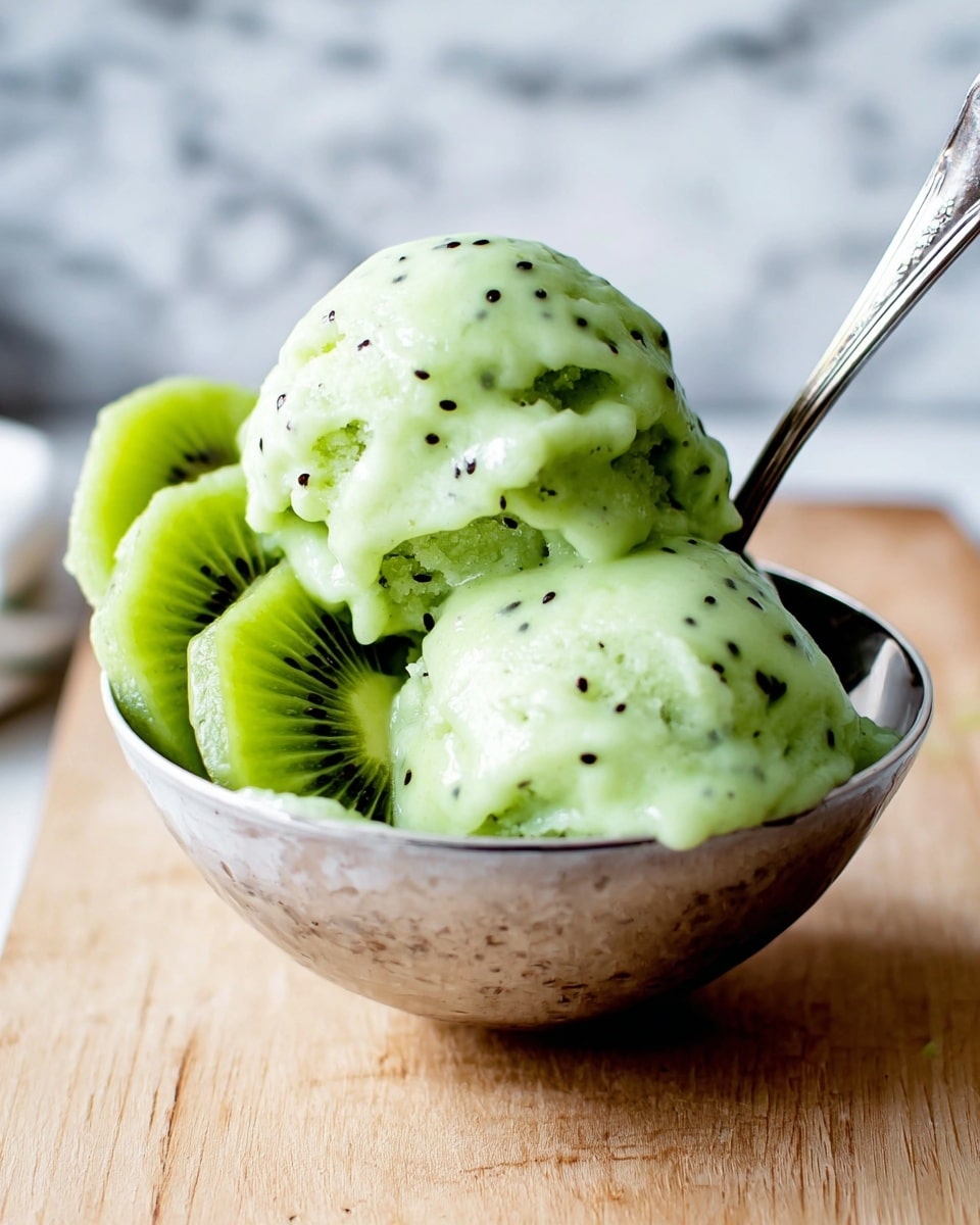 A silver bowl holds two scoops of pale green kiwi sorbet with small black seeds scattered throughout. The sorbet looks smooth and creamy with a soft texture, and there are two slices of fresh kiwi placed beside the scoops inside the bowl. A silver spoon rests inside the bowl, partially covered by the sorbet. The bowl sits on a light wooden surface with a blurred white marbled background. photo taken with an iphone --ar 4:5 --v 7