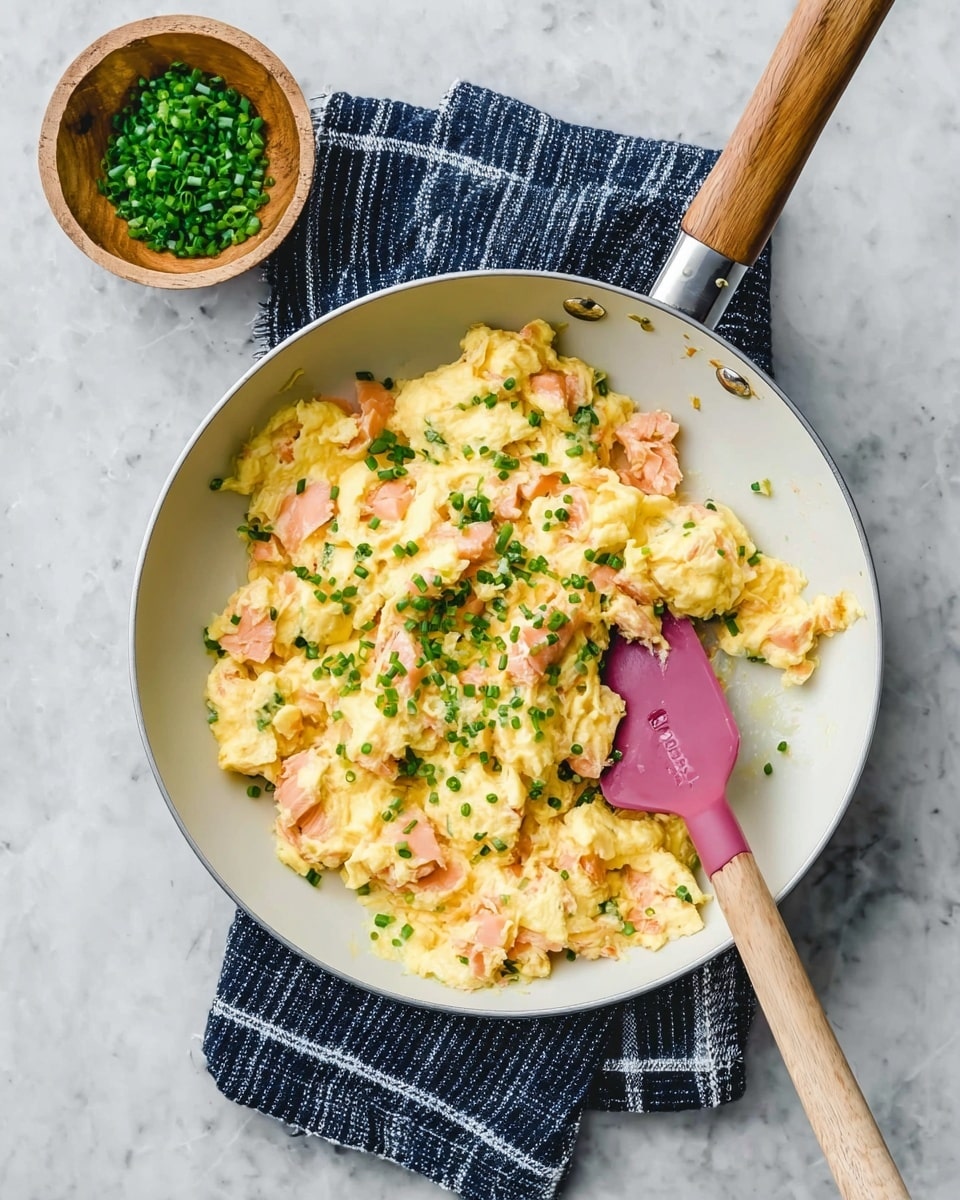 A white pan filled with creamy scrambled eggs mixed with small pieces of pink salmon, topped with small green chive pieces scattered evenly across the dish. A pink spatula with a wooden handle rests inside the pan, which sits on a navy blue cloth with white stripes, all placed on a white marbled surface. A small wooden bowl filled with chopped green chives is positioned next to the pan. Photo taken with an iphone --ar 4:5 --v 7