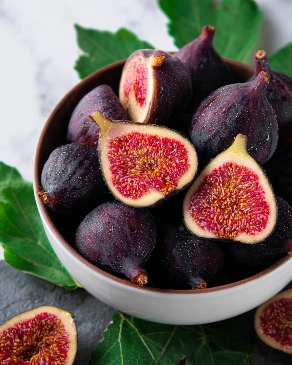 The image shows a white bowl full of whole dark purple figs with a slightly wet look, and some figs cut in half showing a bright reddish-pink inside full of tiny yellow seeds. The cut figs rest on top and around the whole figs, with the inside texture detailed and fresh. The bowl sits on a white marbled surface, and there are a few green fig leaves beside it. Photo taken with an iphone --ar 4:5 --v 7