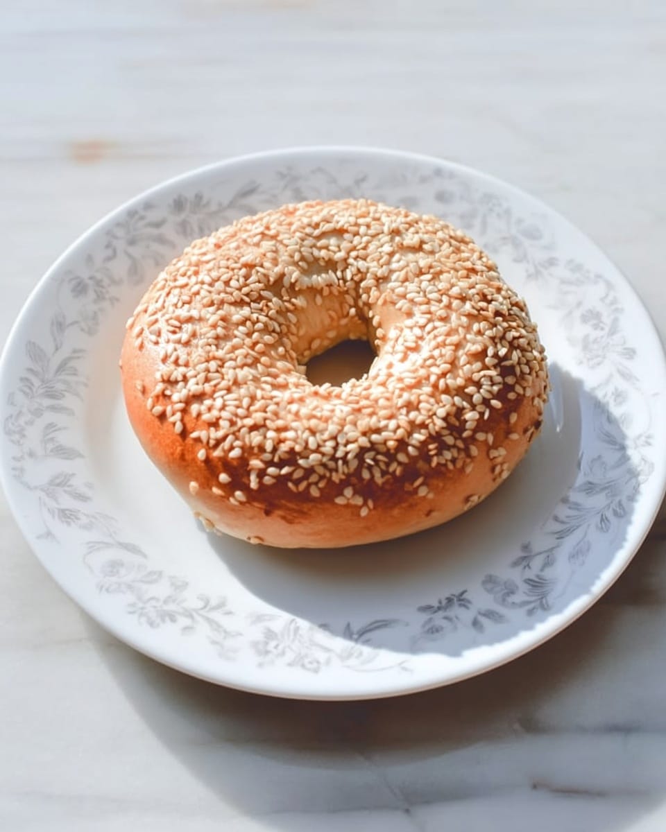 A single golden brown sesame seed bagel sits in the center of a white plate with a subtle floral pattern around the edge. The bagel is round with a smooth, shiny surface topped evenly with pale beige sesame seeds. The bagel’s thickness is moderate, and its hole is round and clear. The plate rests on a white marbled surface with soft natural light casting gentle shadows, highlighting the bagel’s texture and warm color. Photo taken with an iphone --ar 4:5 --v 7