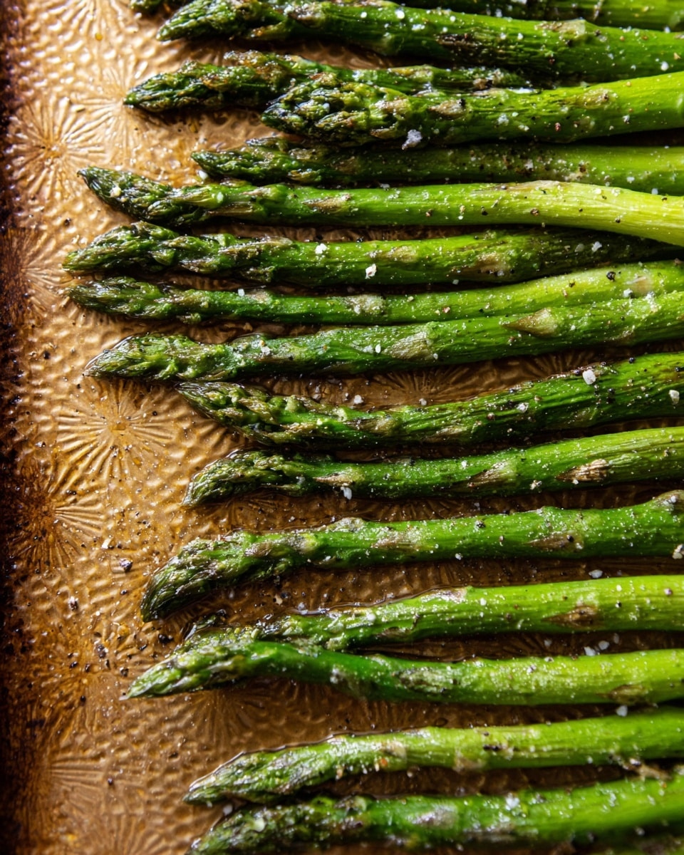 A close-up view of a single layer of roasted green asparagus spears neatly arranged side by side on a textured golden baking tray, sprinkled with coarse salt and cracked black pepper. The asparagus tips are darker green with slight charring, while the stalks show a bright to deep green gradient with a soft, wrinkled texture from roasting. The golden baking tray has small raised patterns and a warm, uneven shine from the cooking process, adding contrast to the vibrant green asparagus. Photo taken with an iphone --ar 4:5 --v 7