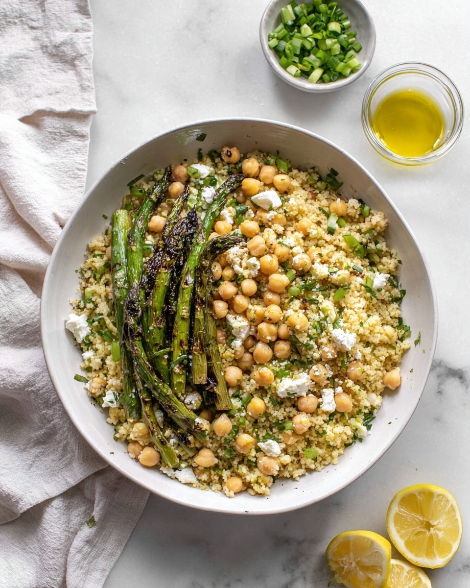 A white bowl filled with three main layers: at the bottom, a layer of small, round, light yellow couscous pearls; mixed throughout, beige chickpeas scattered evenly; on top left, a small bunch of grilled green asparagus spears with char marks. The couscous and chickpeas mix also has small pieces of white cheese and chopped green herbs. The bowl sits on a white marbled surface with a light cloth folded on the top left, a small white bowl of chopped green onions at the top right, a small glass bowl of yellow olive oil with herbs just below that, and two lemon halves with juice spilled around on the bottom right. Photo taken with an iphone --ar 4:5 --v 7