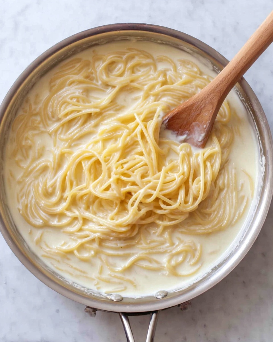 Inside a silver pan on a white marbled surface, there is a creamy sauce with cooked spaghetti noodles mixed in it. The noodles are pale yellow and soft, floating in a thick, white creamy liquid that covers them evenly. A wooden spoon is resting inside the pan, with its handle stretching out toward the right side of the image, partially submerged in the sauce and noodles. The texture of the sauce looks smooth and rich, surrounding the strands of pasta gently. Photo taken with an iphone --ar 4:5 --v 7