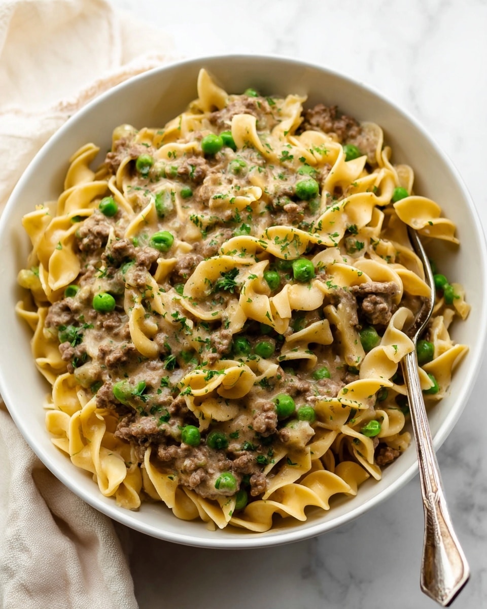 A white bowl filled with three layers of creamy beef and noodles. The bottom layer is wide, curly noodles in a light yellow color. The middle layer is small pieces of cooked brown ground beef mixed with bright green peas scattered evenly. The top layer has more noodles, beef, and peas covered in a creamy sauce with green herbs sprinkled over the dish. On the top right inside the bowl, there is a silver fork resting in the food. The bowl is placed on a white marbled surface with a soft cloth in the background. Photo taken with an iphone --ar 4:5 --v 7