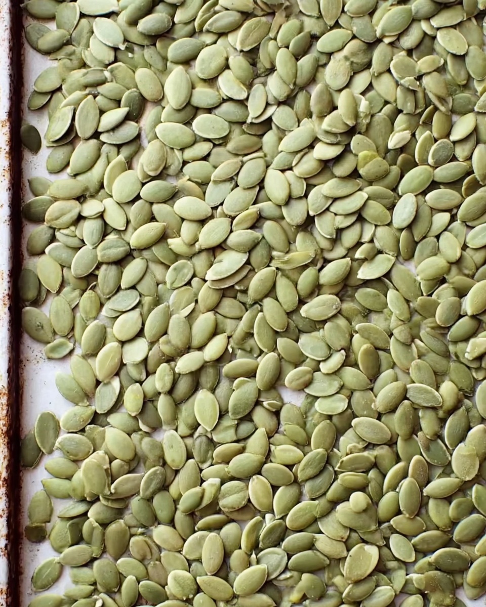 The image shows a close-up of a flat baking tray filled evenly with many small, flat, pale green pumpkin seeds that cover the entire surface. The seeds are smooth and slightly shiny with a gentle curve, laid out in a single thin layer filling every part of the rectangular tray. The background is a white marbled texture. Photo taken with an iphone --ar 4:5 --v 7