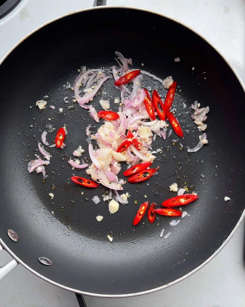 Inside a black non-stick frying pan on a white marbled surface, there are small uneven pieces of white garlic scattered around. Thin, pale purple slices of shallots are spread mostly on the left and upper areas, mixed lightly with the garlic. Bright red sliced chili peppers, some whole and some cut, are placed scattered evenly around the pan. There is a light coating of oil visible, especially in the center, giving a slight shine to the ingredients. The edges of the pan are silver, and the background includes parts of a white stovetop. photo taken with an iphone --ar 4:5 --v 7