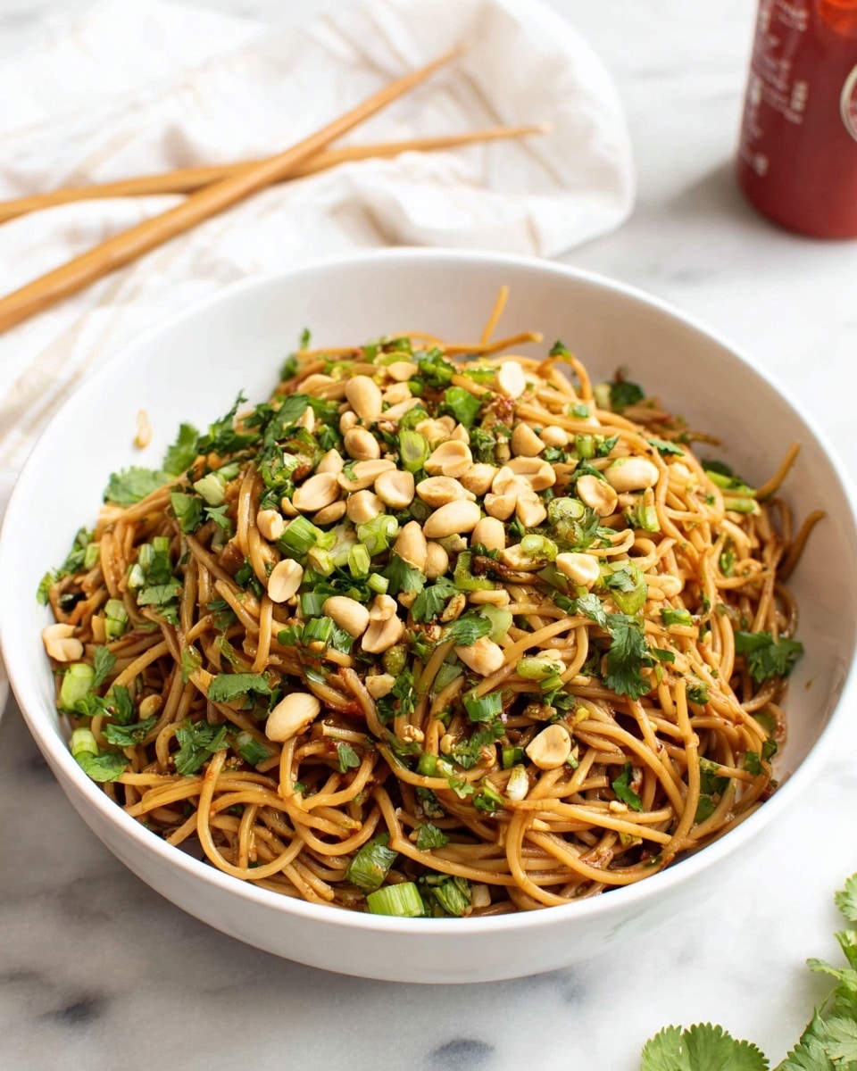 A white bowl filled with a heap of thin brown noodles, mixed with chopped green onions and fresh cilantro leaves scattered throughout. Light golden peanuts are spread on top, adding a crunchy texture. The noodles look shiny with a light sauce coating. The bowl is placed on a white marbled surface, with a pair of wooden chopsticks resting on a white cloth in the background and a red sauce bottle partially visible. photo taken with an iphone --ar 4:5 --v 7