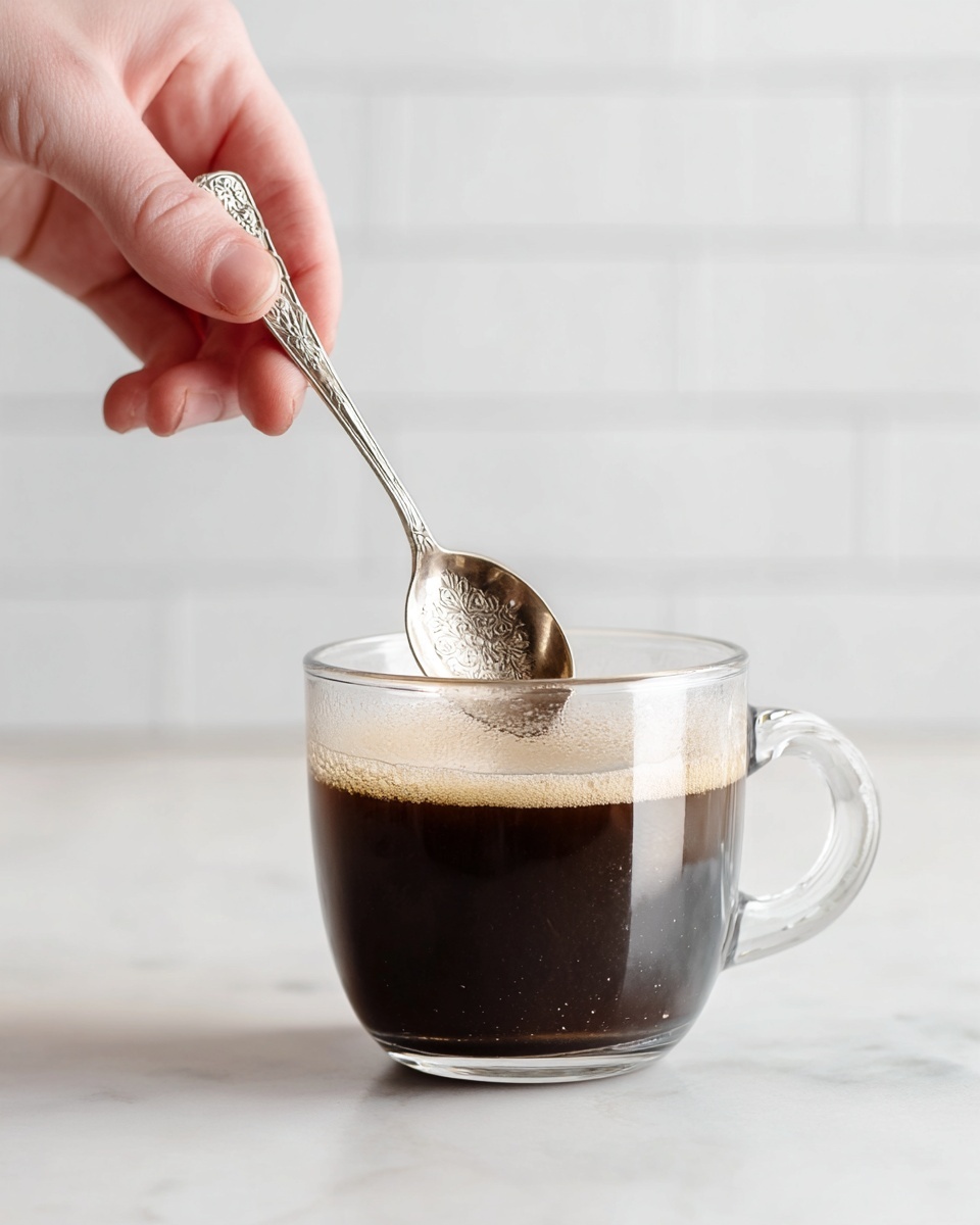 A clear glass cup filled halfway with dark brown coffee sits on a white marbled surface. A woman's hand holds a detailed silver spoon, stirring the coffee inside the cup. The cup has a curved handle on one side and light reflections from nearby light sources shimmer on the glass and liquid. In the background, a softly focused white tile wall adds to the simple, clean look. photo taken with an iphone --ar 4:5 --v 7