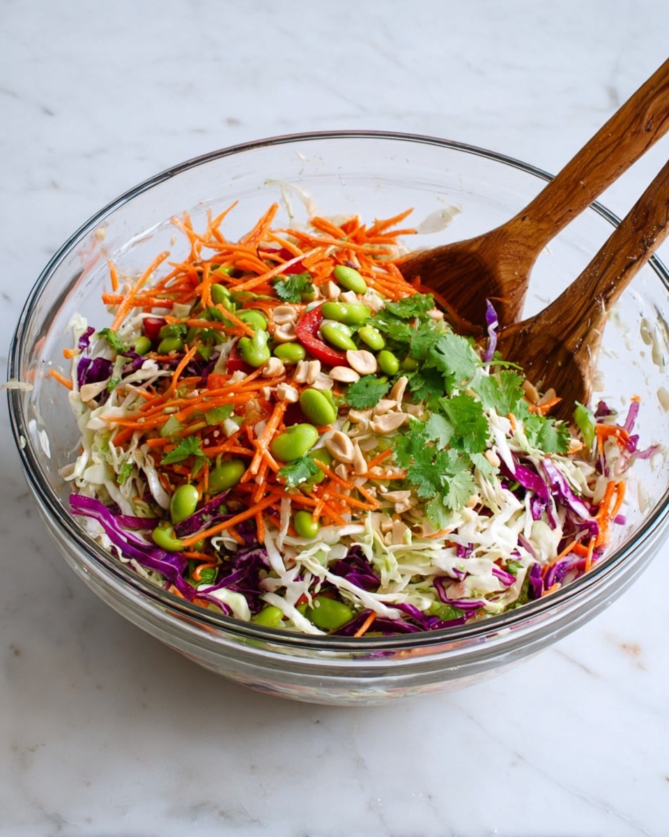 A clear glass bowl filled with a colorful mixed salad sits on a white marbled surface. The salad has multiple layers: shredded white cabbage at the bottom, thin strips of orange carrots and purple cabbage mixed throughout, bright green edamame beans and fresh green cilantro leaves scattered on top, along with pieces of red bell pepper and a few peanuts adding texture and color. Two wooden spoons rest in the bowl from opposite sides, partially covered with the salad. photo taken with an iphone --ar 4:5 --v 7