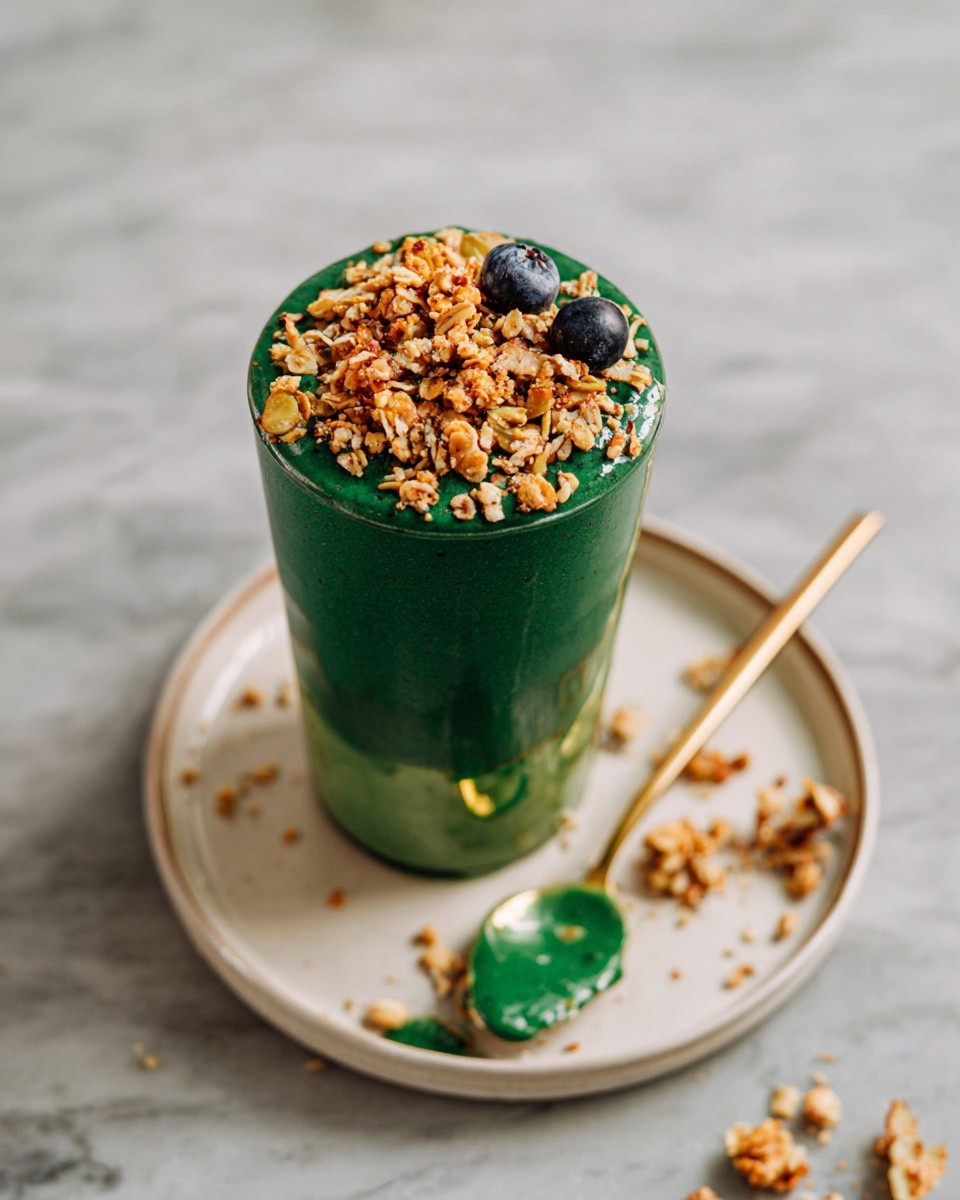 A tall clear glass is filled with two thick layers of dark green smoothie, topped with a generous layer of golden brown granola and a few dark blueberries on the top right. The glass sits on a white plate with a gold spoon resting inside, showing some smoothie on the spoon. Some granola pieces and blueberries are scattered on the white marbled surface around the plate. The overall look is fresh and natural with highlights on the green and golden colors, photo taken with an iphone --ar 4:5 --v 7