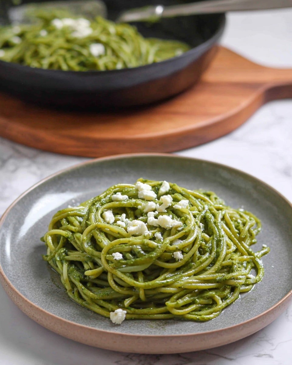 A plate with one layer of green spaghetti coated in a smooth green pesto sauce sits in the foreground, topped with small white cheese crumbles scattered on top. Behind it, a black pan filled with more green pesto spaghetti is partially visible, resting on a wooden board. The whole scene is set on a white marbled surface. photo taken with an iphone --ar 4:5 --v 7