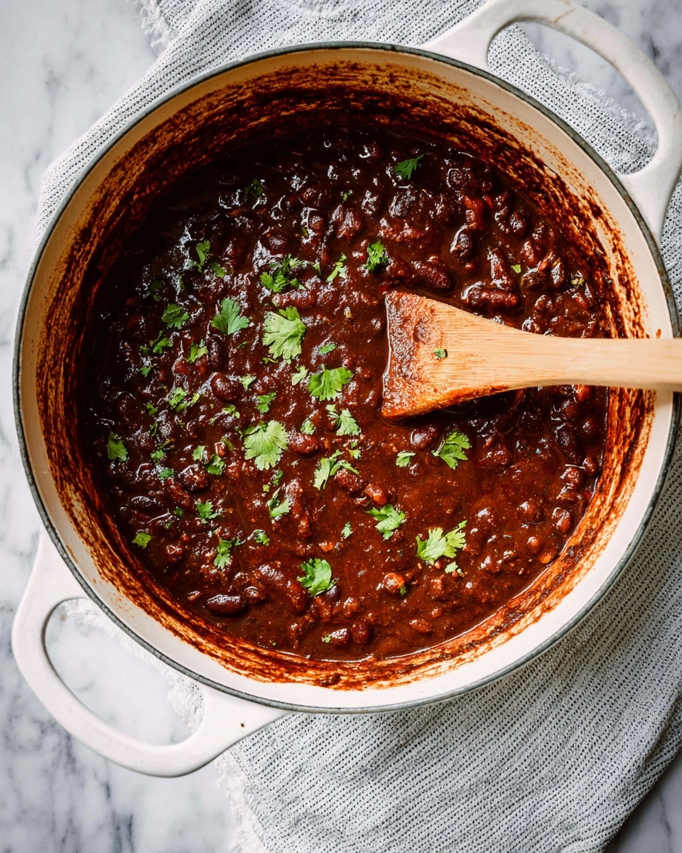 The image shows a white pot filled with thick, dark brown chili with visible chunks and beans throughout the rich sauce. Small green cilantro leaves are sprinkled on top, adding a fresh contrast to the deep brown color. A wooden spoon with a light wood handle is resting inside the pot, partially covered with the chili. The pot sits on a white marbled surface with a light gray striped cloth below the pot handle. The edges of the pot have some chili stains, showing it has been cooked and stirred well. photo taken with an iphone --ar 4:5 --v 7