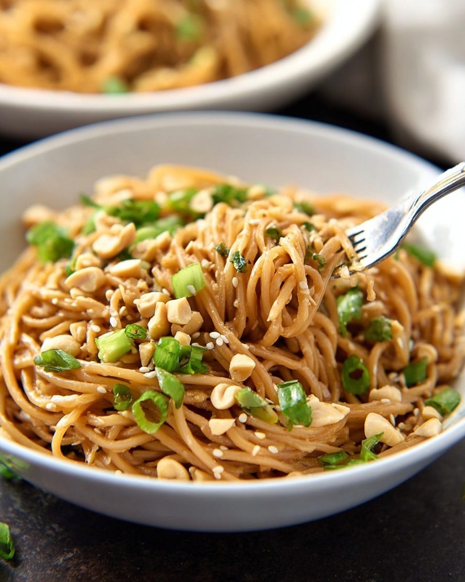 A white bowl filled with cooked brown noodles mixed with light brown sauce, sprinkled generously with small white sesame seeds and light tan chopped peanuts, and garnished with bright green chopped scallions scattered throughout. A silver fork is twirling some noodles, lifting them from the bowl. The bowl sits on a dark surface with a blurred white bowl of more noodles in the background. Photo taken with an iphone --ar 4:5 --v 7