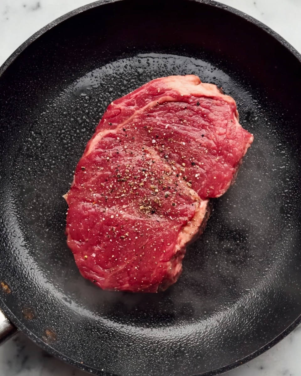 A single raw steak with a red color and some white fat edges is placed in the center of a black skillet. The steak is sprinkled with black pepper and salt, showing a slight texture of marbled fat within the meat. The skillet has a shiny, smooth surface with a little steam rising near the bottom edge of the steak, suggesting it's just starting to cook. The background is a white marbled surface. photo taken with an iphone --ar 4:5 --v 7