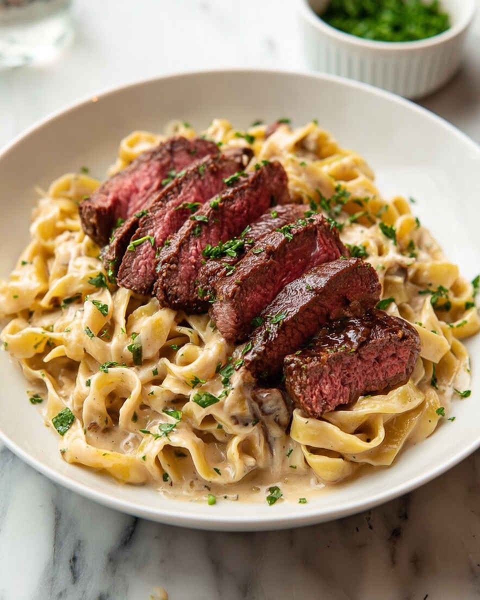 A white round plate holds a serving of thick, creamy pasta at the bottom layer, with flat, wide noodles coated in a light beige sauce. On top, there are several slices of medium-rare steak arranged neatly in a row, showing a warm pink center and a browned, seared crust. The dish is sprinkled with chopped green herbs scattered over both the pasta and steak. The plate sits on a white marbled surface, and a small white bowl with green garnish is blurred in the background. Photo taken with an iphone --ar 4:5 --v 7